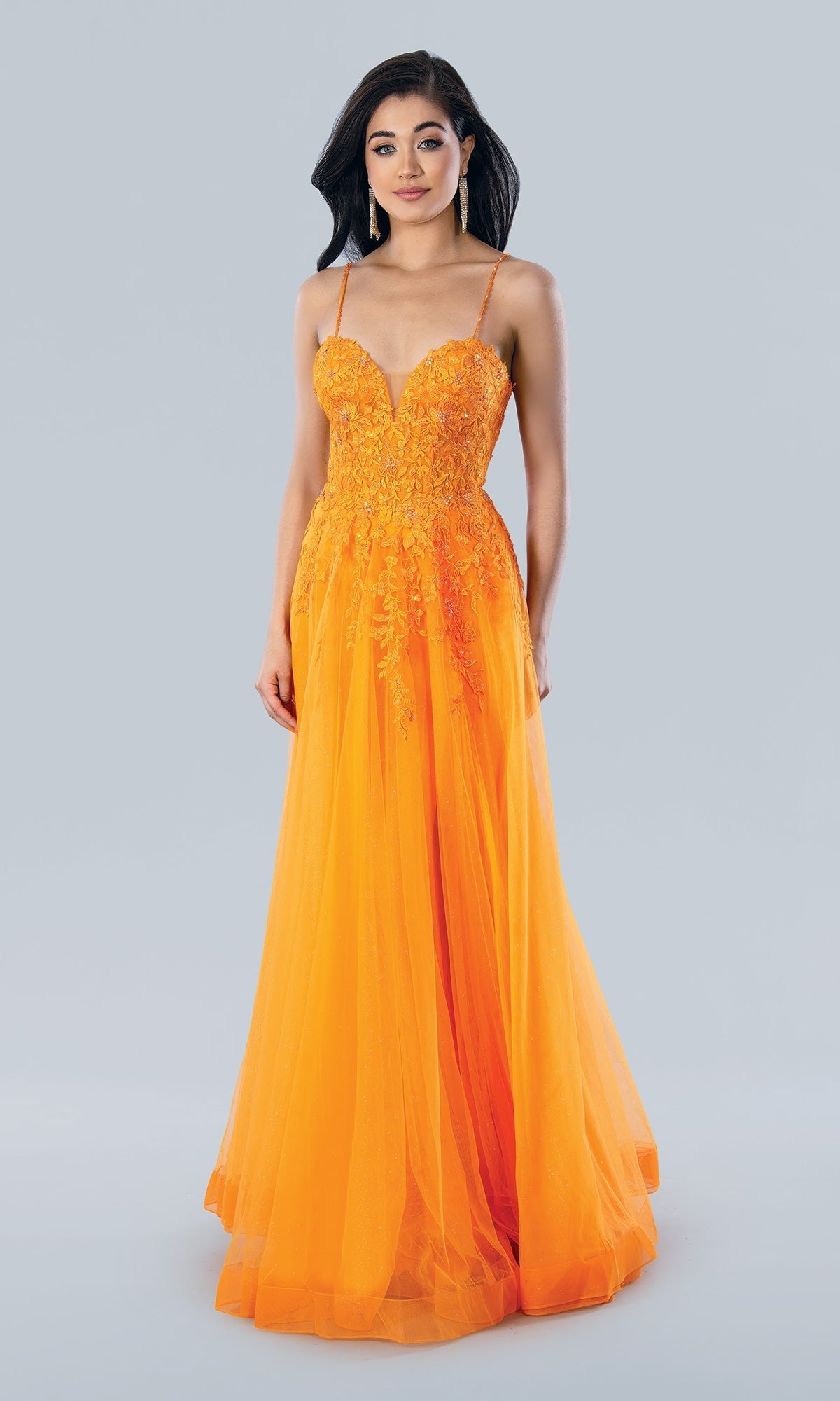 A woman stands against a plain light background wearing the Stella Couture 22027, a floor-length bright orange A-line prom dress featuring thin straps and a lace embroidered bodice.