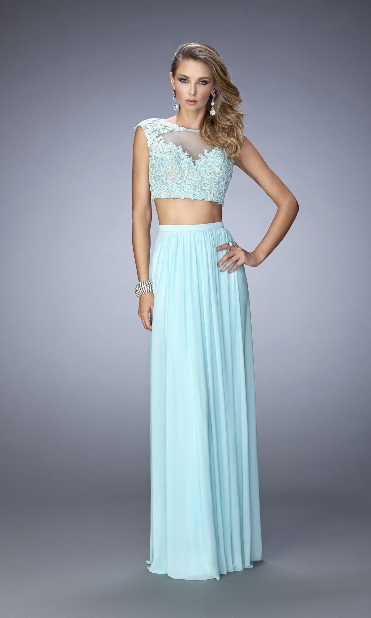 A woman wears the La Femme Two-Piece Lace-Top Jersey Prom Dress 22110, featuring a light blue lace crop top and a long, flowing skirt, standing against a plain gray background.