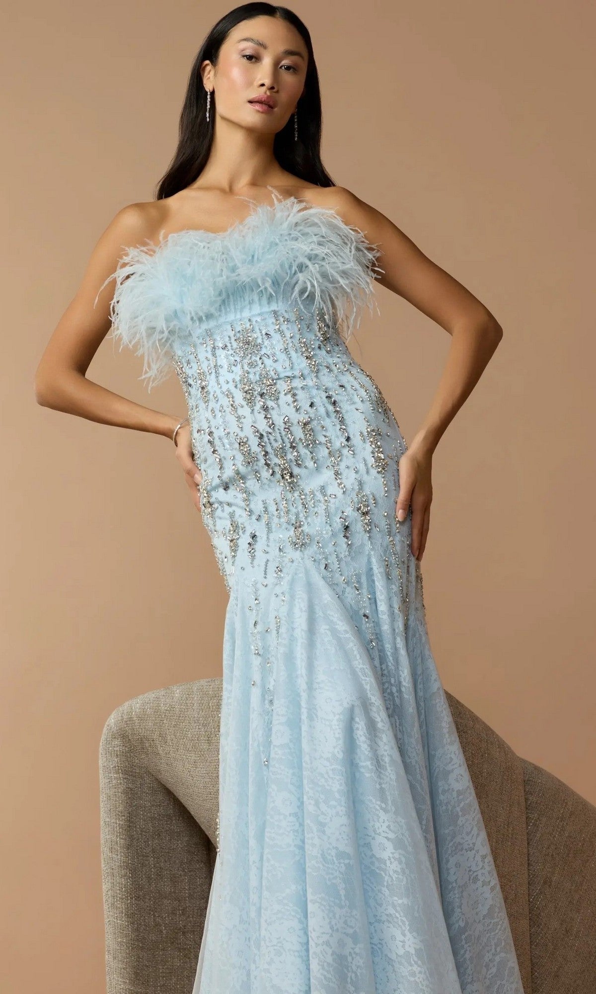 A woman stands against a beige background wearing the Mac Duggal 2238 long formal dress, a strapless designer gown featuring a feather-embellished neckline and silver accents.