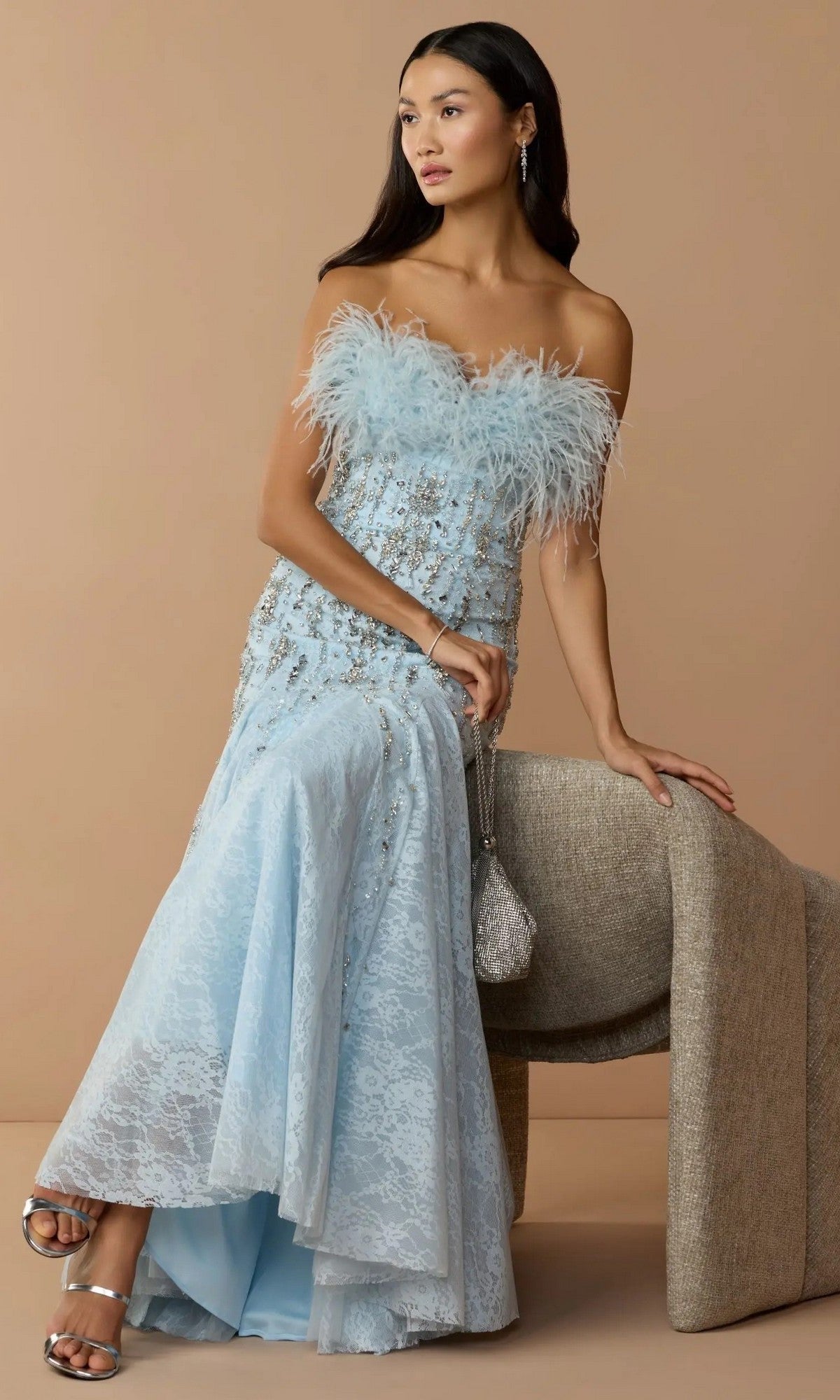 A woman in the Mac Duggal 2238 Long Formal Dress, a light blue strapless gown with a lace and feather-embellished neckline, sits on a beige chair against a tan background.