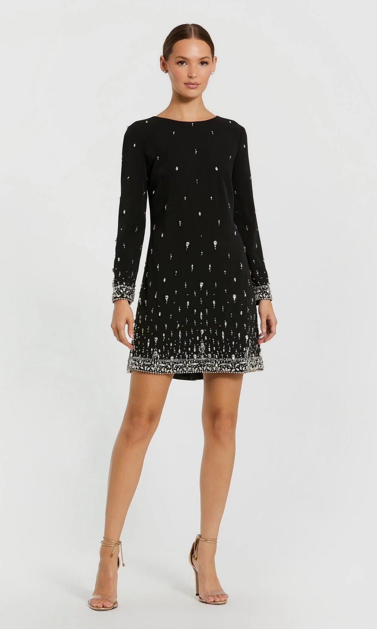 A woman stands with one hand on her hip and a neutral expression, wearing the Mac Duggal 2262 Short Semi-Formal Dress—a black, long-sleeve mini dress with crystal beading.