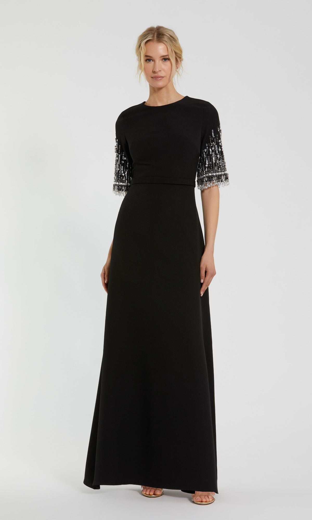 A woman wears the Mac Duggal 2270 Short Sleeve Sleek Formal Dress, a floor-length black gown with embellished short sleeves, styled with neutral sandals against a plain background.