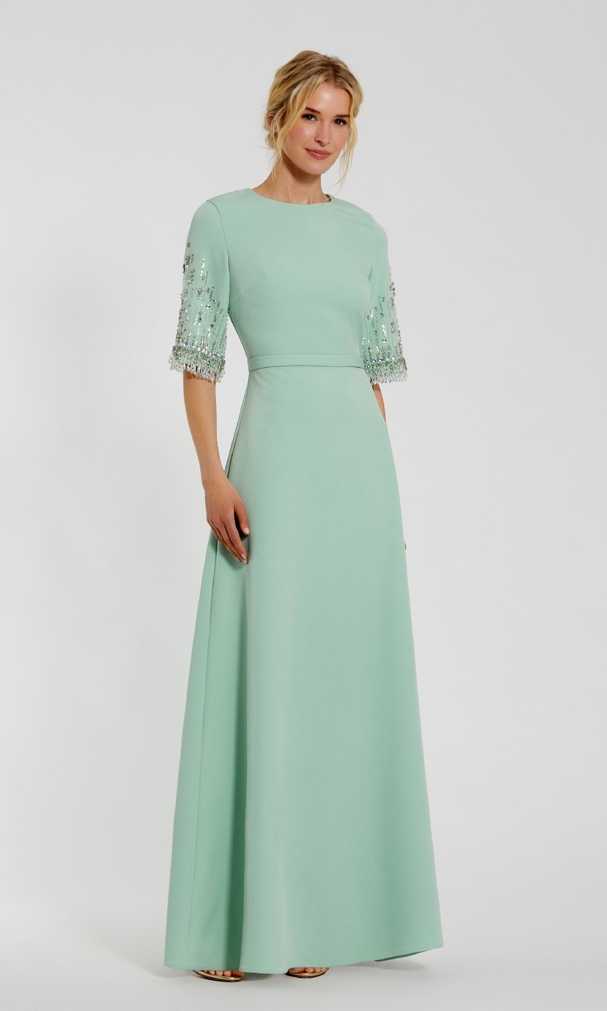A woman in a light green Mac Duggal 2270 Short Sleeve Sleek Formal Dress, featuring sparkling bead embellishments, stands against a white background.