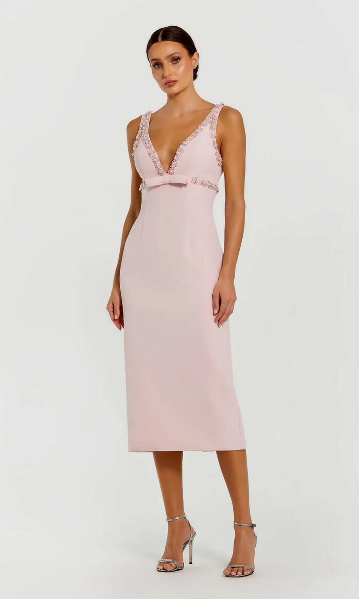 A woman wears the Mac Duggal 2276 V-Neck Midi Wedding Guest Dress in blush with lace trim, a front bow, and silver heels—an elegant semi-formal look. She poses against a plain background.