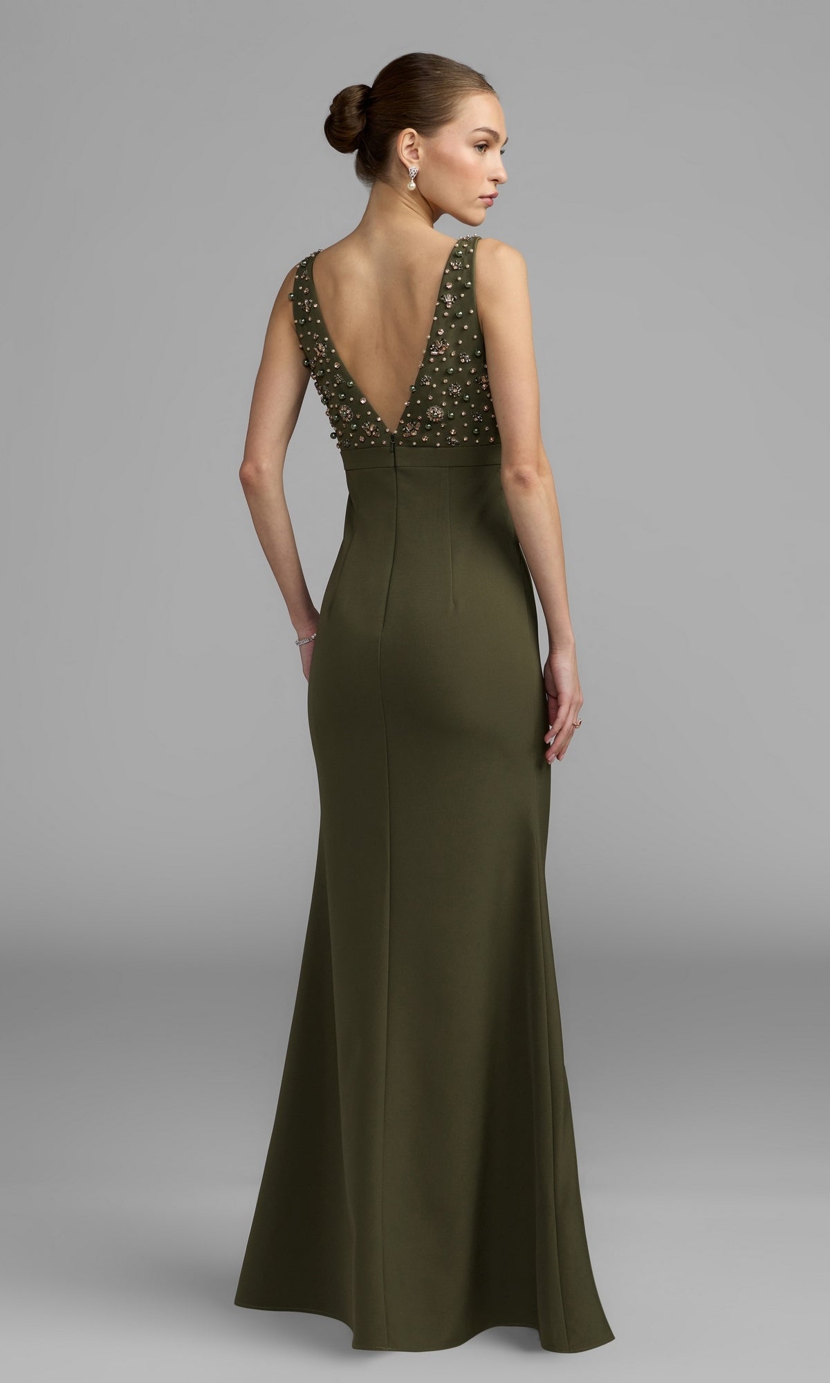 A woman wearing the Mac Duggal 2278 Beaded-Bodice Long Prom Dress in olive green, featuring an empire waist, deep V-back, and intricately beaded bodice, stands against a plain gray background.