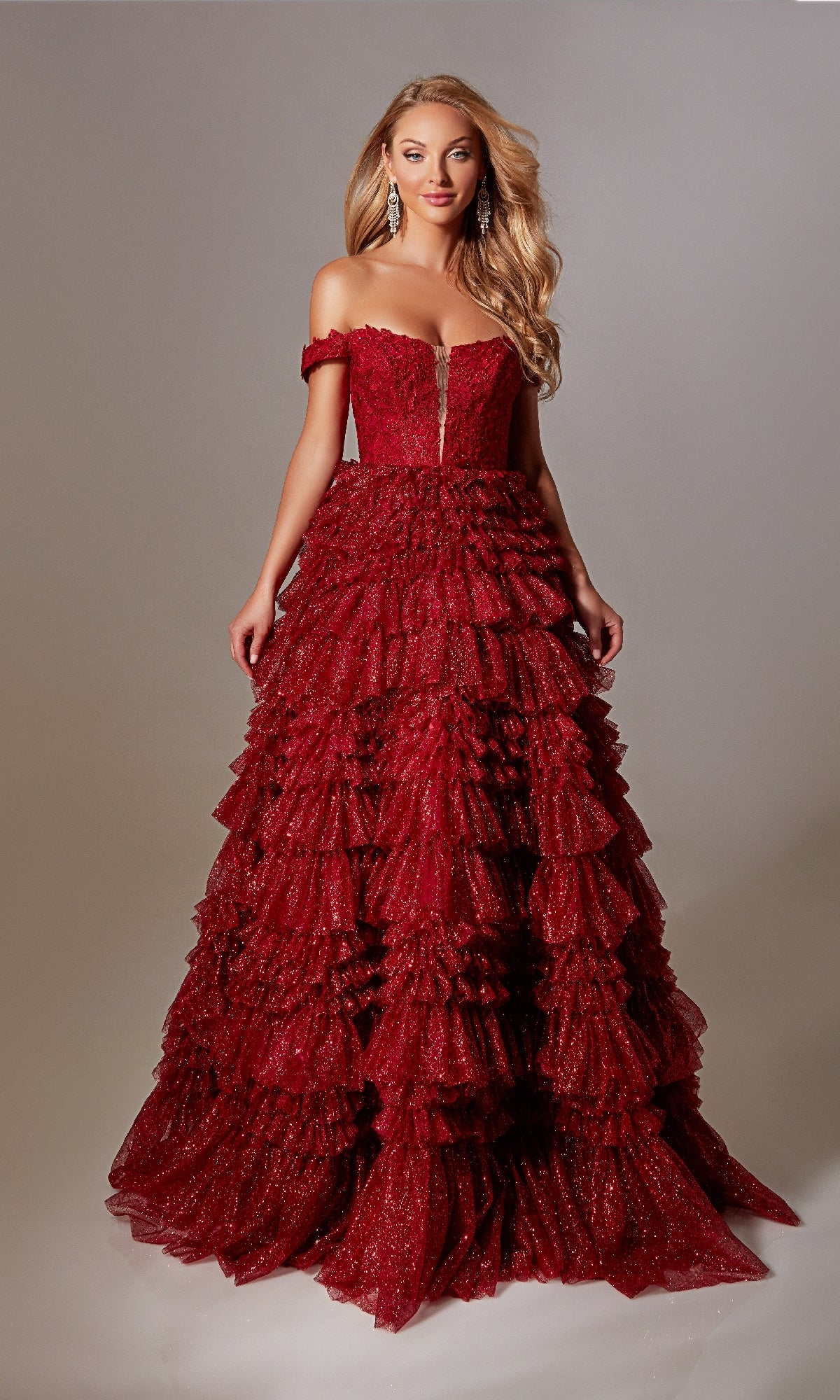 A woman wears the Lace-Up Long A-Line Glitter Prom Dress with Ruffles, featuring red sparkly details and layered ruffles, as she stands against a plain gray background.