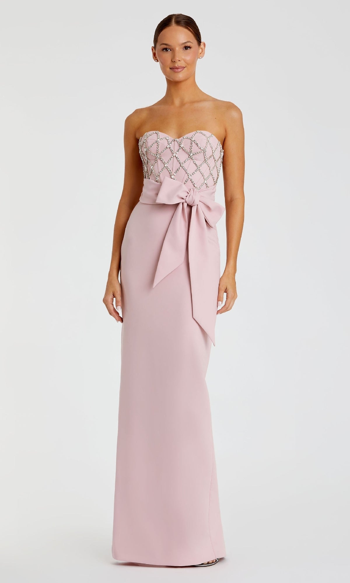 A woman models the Mac Duggal 2294 Strapless Sweetheart Formal Gown in blush pink with a beaded bodice and large waist bow, standing against a plain white background.