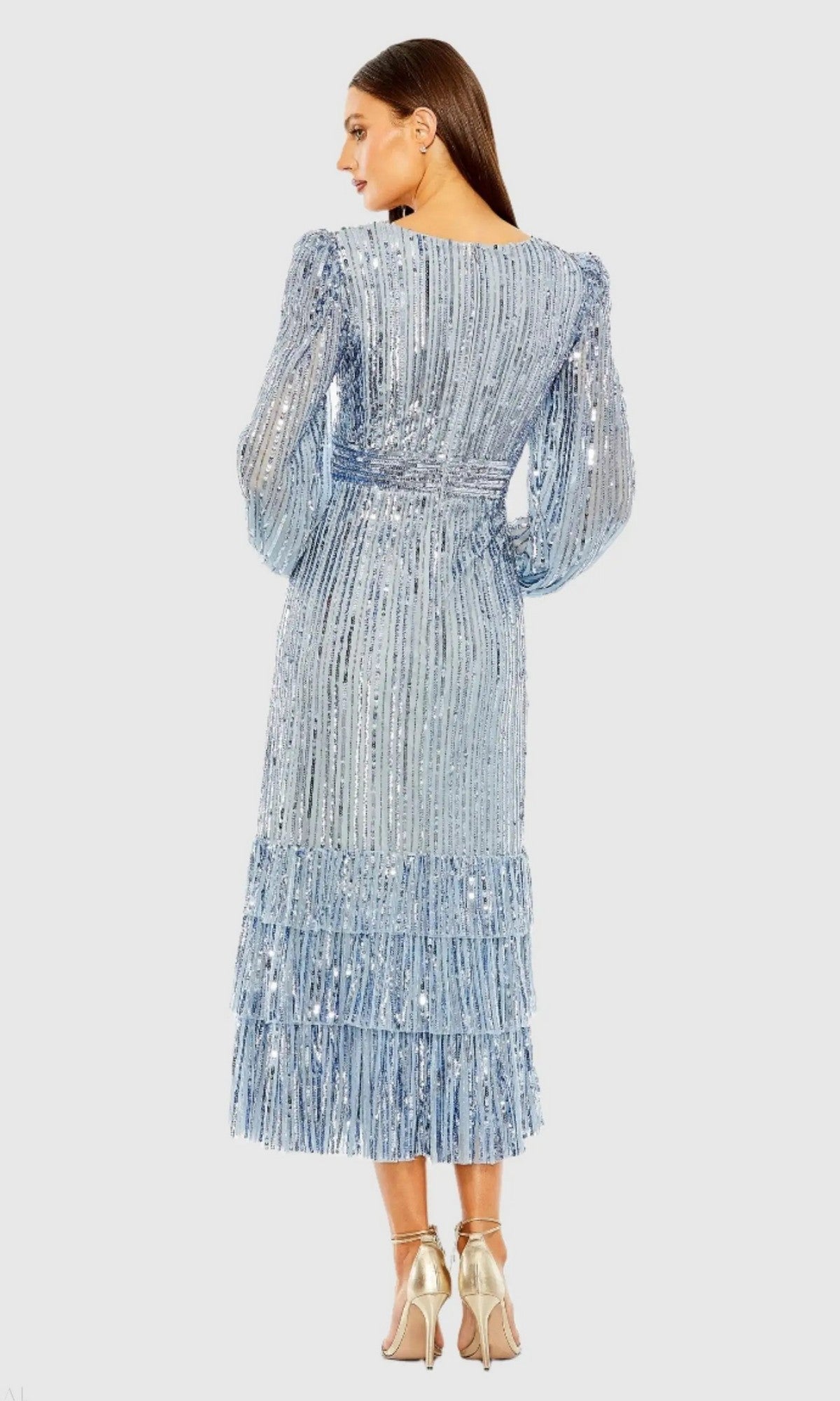 A woman stands facing away, wearing the Mac Duggal 23006 midi semi-formal dress in light blue sequins with tiered layers, a subtle v-neckline, paired with silver high-heeled sandals.