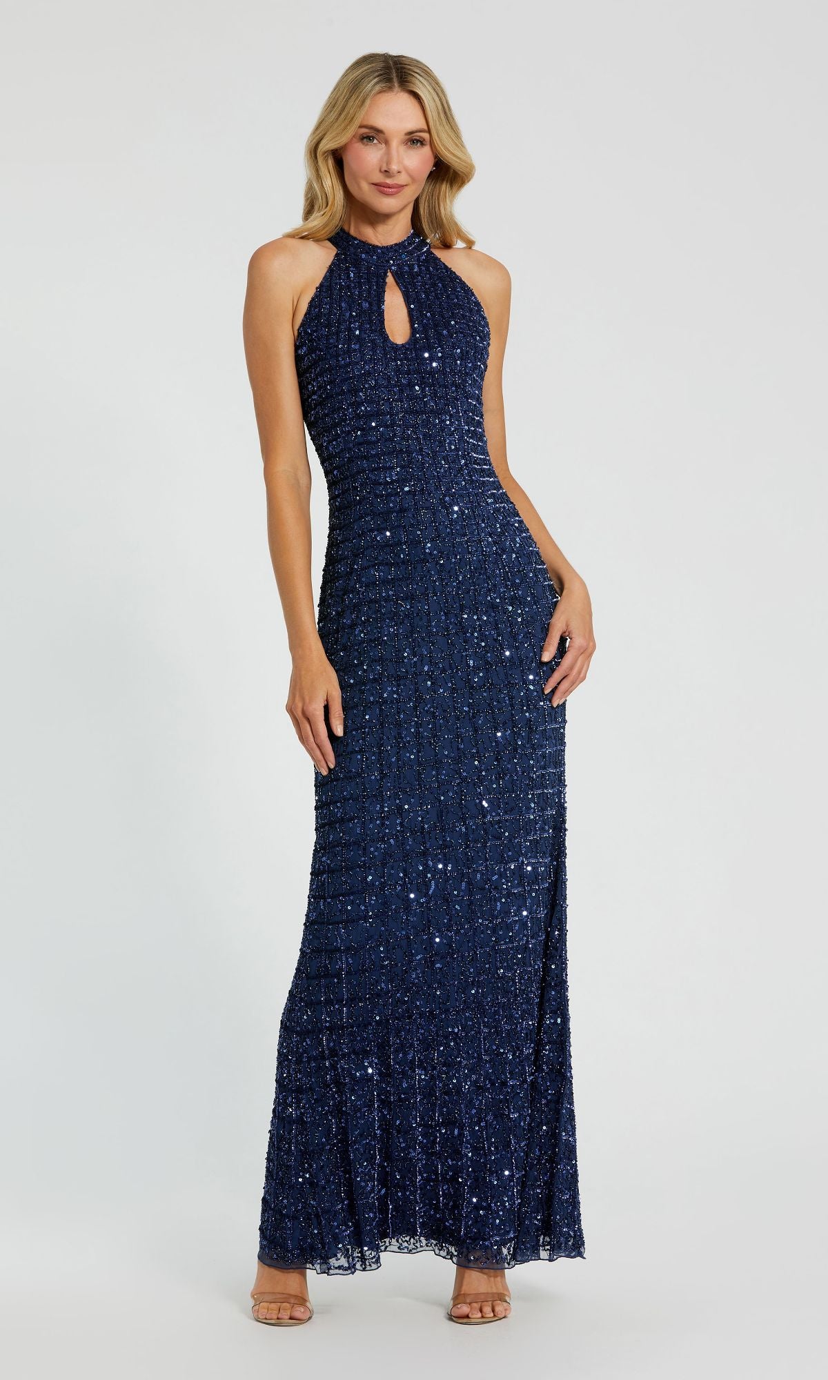 A person wearing the Mac Duggal 23018 High-Neck Long Beaded Prom Dress, a sleeveless navy evening gown with a keyhole neckline, styled with nude heels and posing against a plain light background.