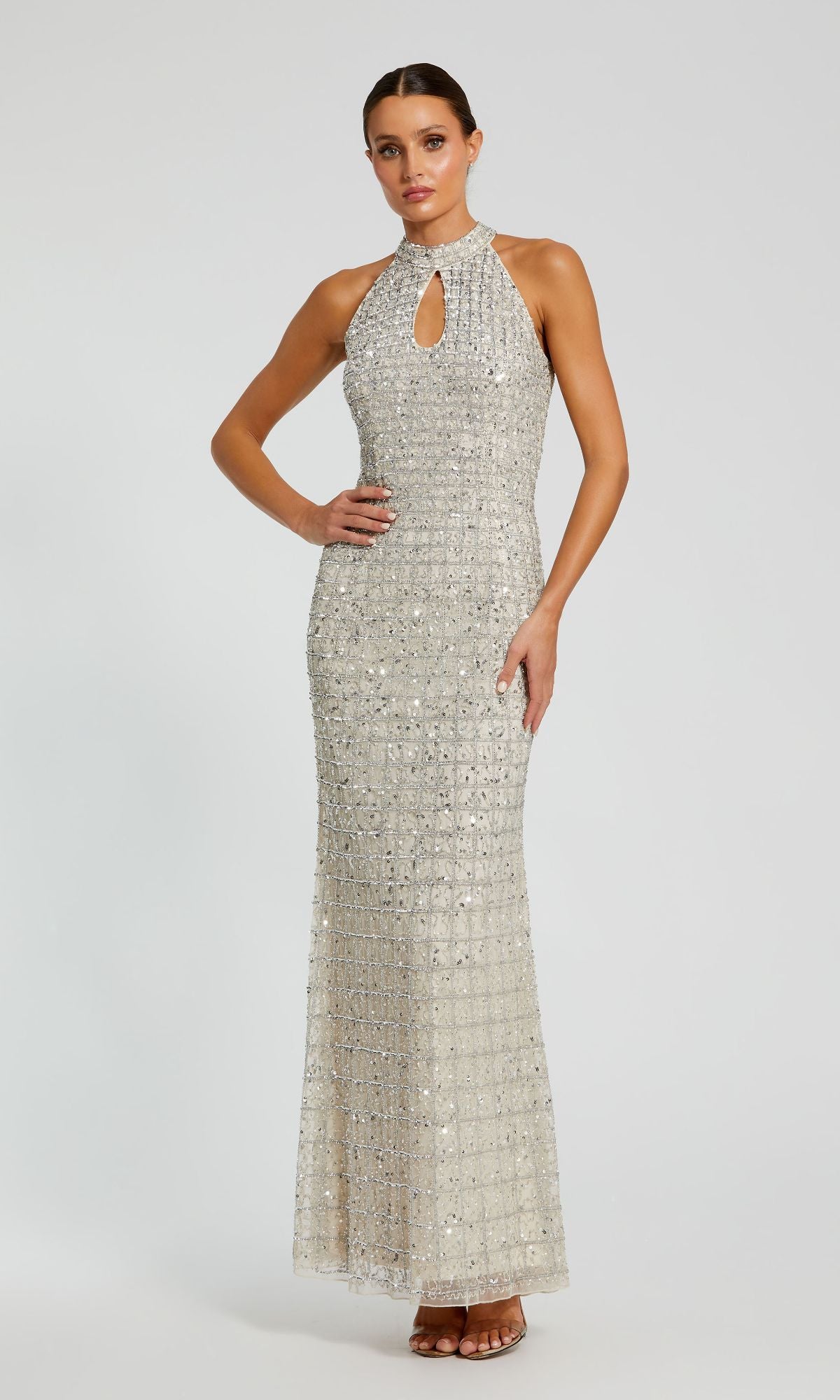 A woman poses against a plain light background in the Mac Duggal 23018: a sleeveless, floor-length, silver beaded prom dress with a high neckline and fitted silhouette.