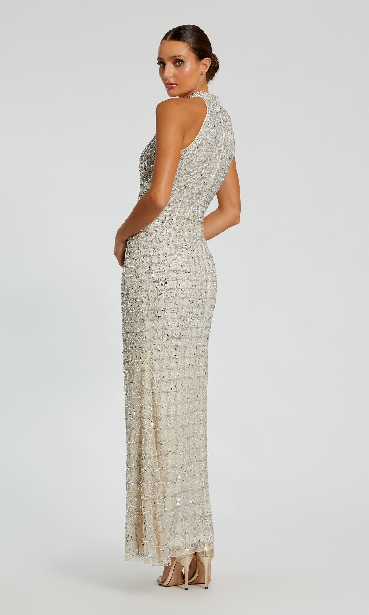 Woman wearing the High-Neck Long Beaded Prom Dress by Mac Duggal 23018, a sleeveless, floor-length gown sparkling with silver sequins, poses gracefully against a white background for an elegant evening look.