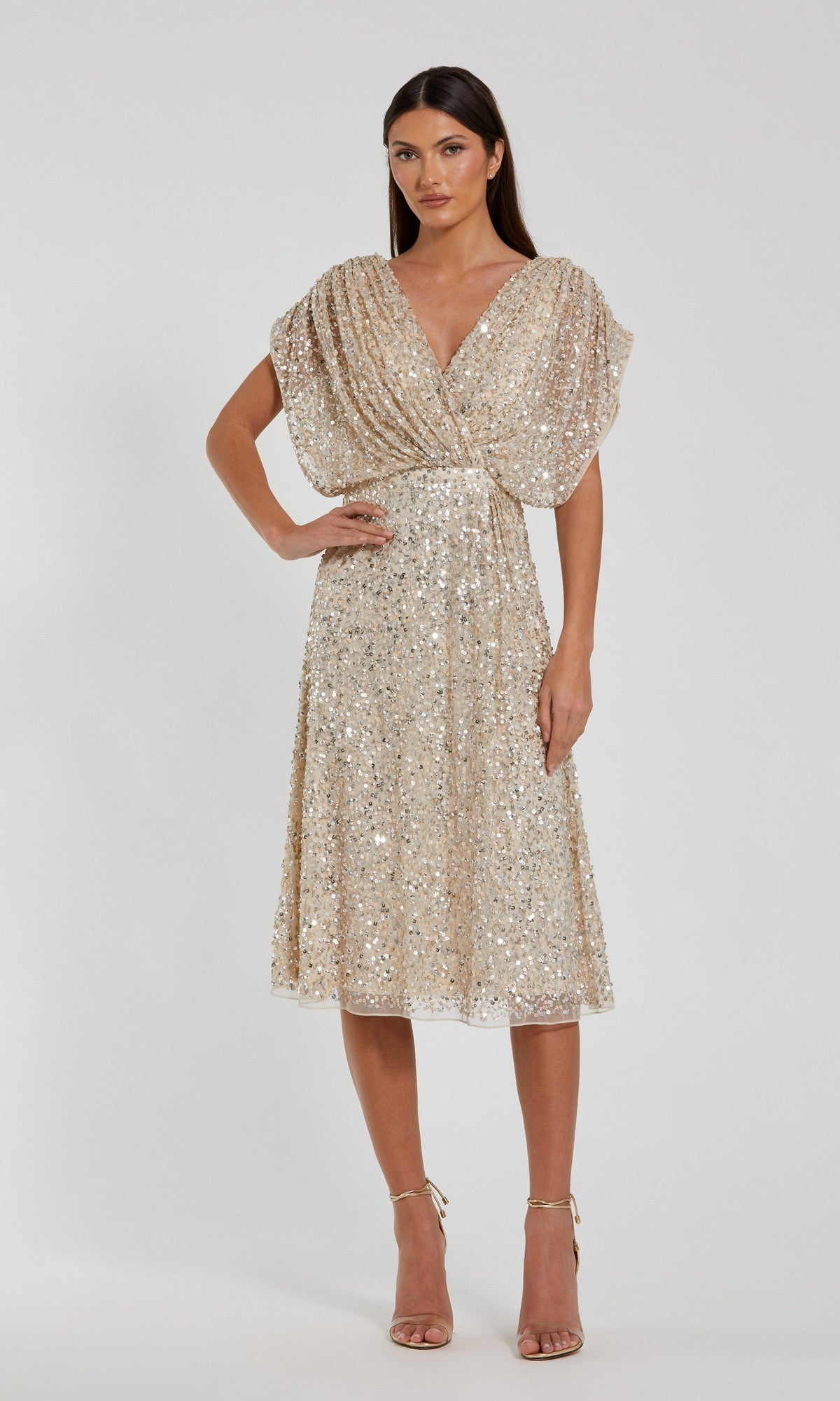 A woman stands against a plain white background wearing the Mac Duggal 23027 Knee Length Party Dress in Nude Silver, featuring short sleeves, sparkling sequins, a flattering v-neck bodice, and matching ankle-strap heels.