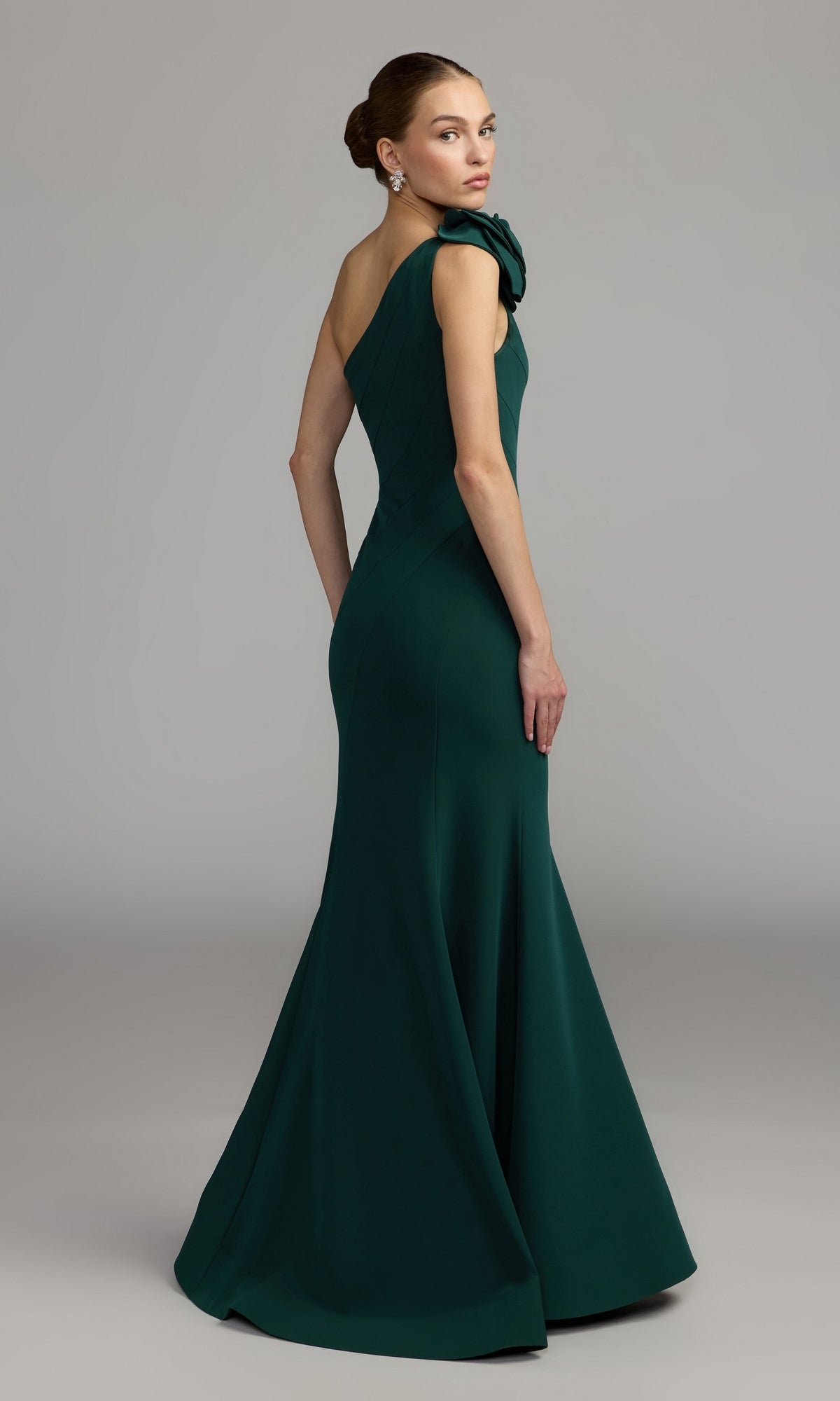 A woman stands against a plain gray background wearing the Mac Duggal 2304 Flower One-Shoulder Formal Dress—a floor-length, dark green mermaid gown featuring a fitted bodice and flared skirt.