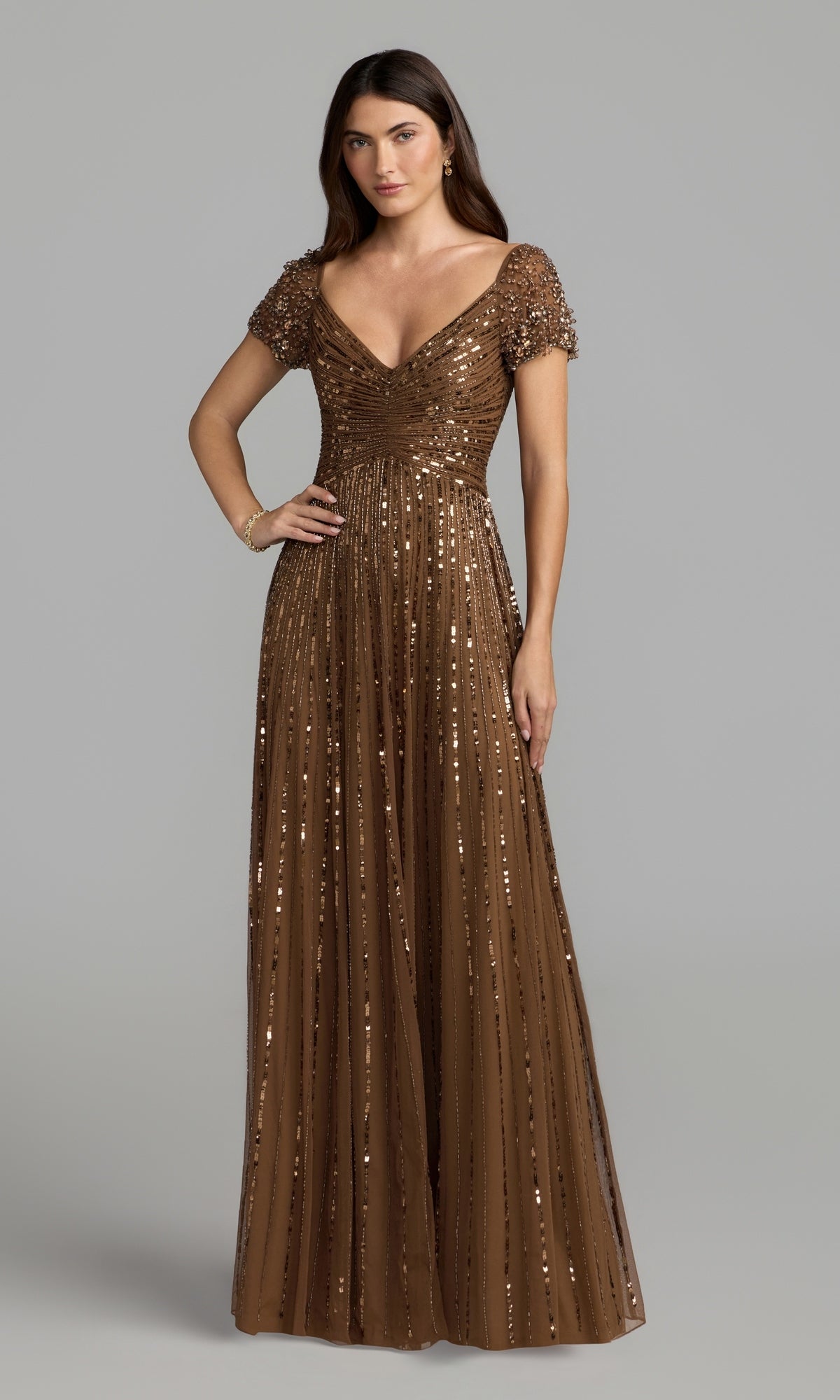Wearing the Mac Duggal 23042 Short Sleeve Sequin Formal Dress in a rich chocolate hue, a woman stands against a plain gray background. This floor-length gown features shimmering sequins, short sleeves, and a V-neckline, making it an elegant choice for any sophisticated event or as a timeless mother of the bride dress.
