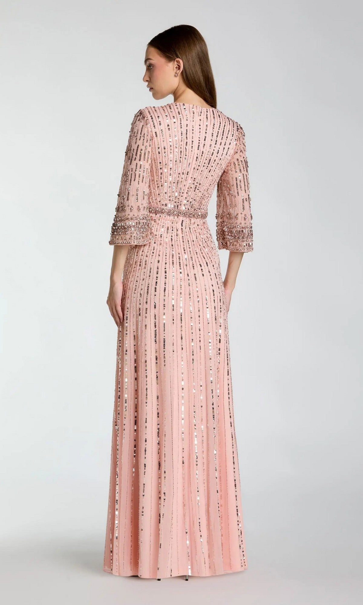 A woman stands facing away, wearing the Mac Duggal 23045 Long Formal Dress in pale pink. This modest evening gown features elbow-length sleeves, a fitted waist, and vertical lines of sequin embellishments.
