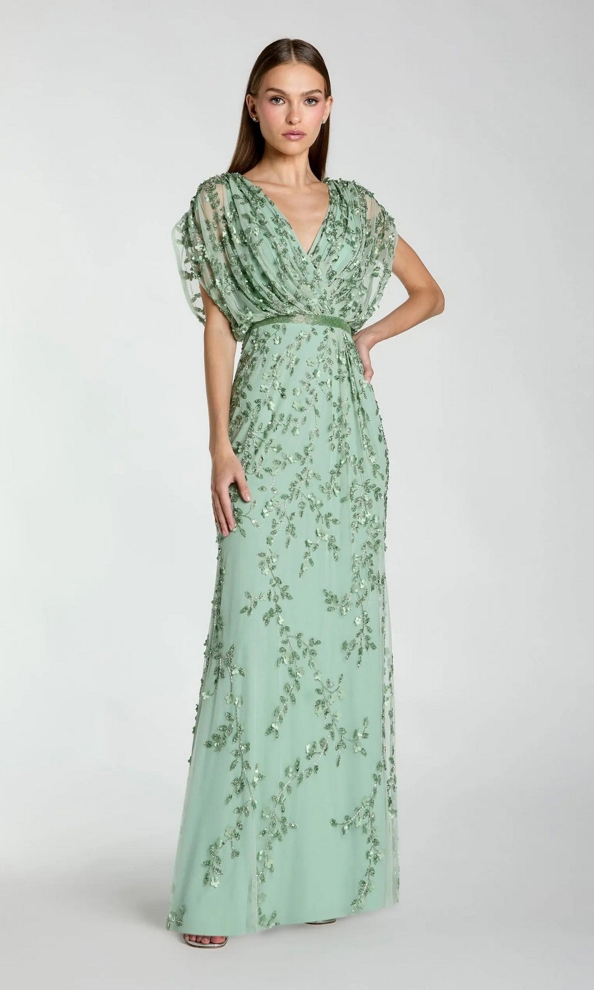 A woman poses with one hand on her hip against a plain background, wearing the Long Formal Dress: Mac Duggal 23067 in seafoam—a floor-length gown featuring short, sheer sleeves and exquisite floral embroidery.