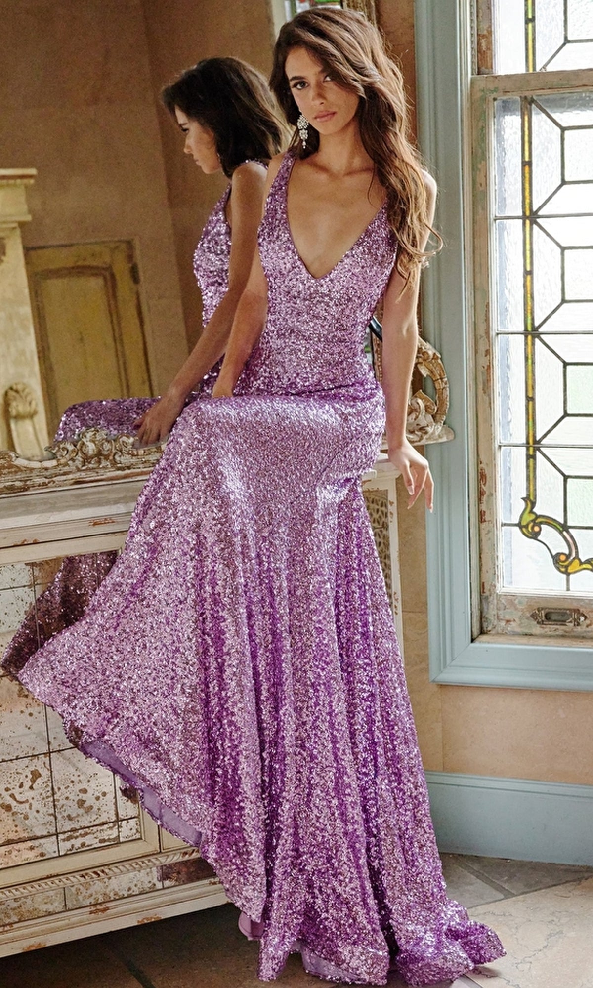 A woman sits on a marble countertop near a stained glass window, her purple Jovani Long Sequin Mermaid Prom Dress 23079 shimmering in the light.