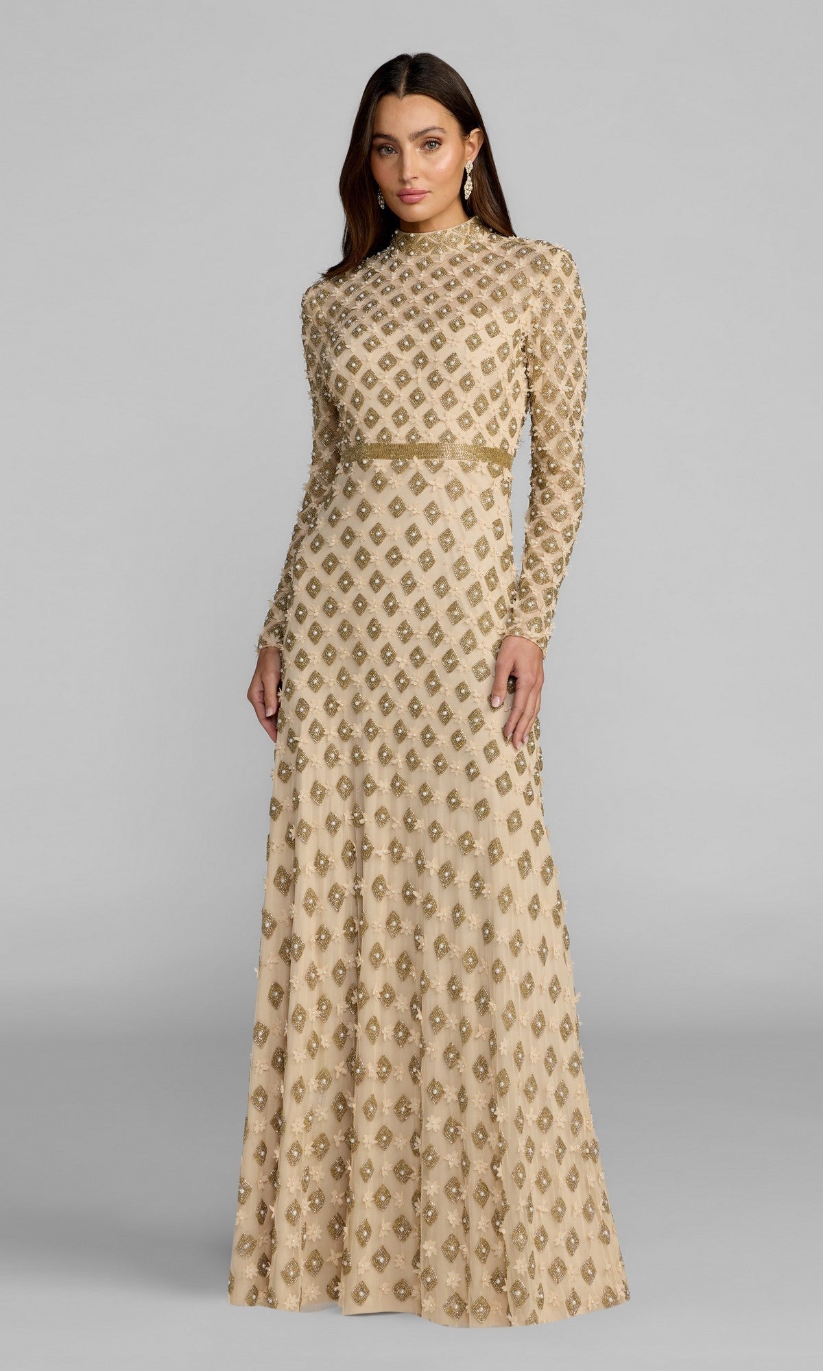 A woman wears the Mac Duggal 23087 long formal dress in champagne gold, featuring long sleeves and embellished patterns, as she stands against a plain gray background.