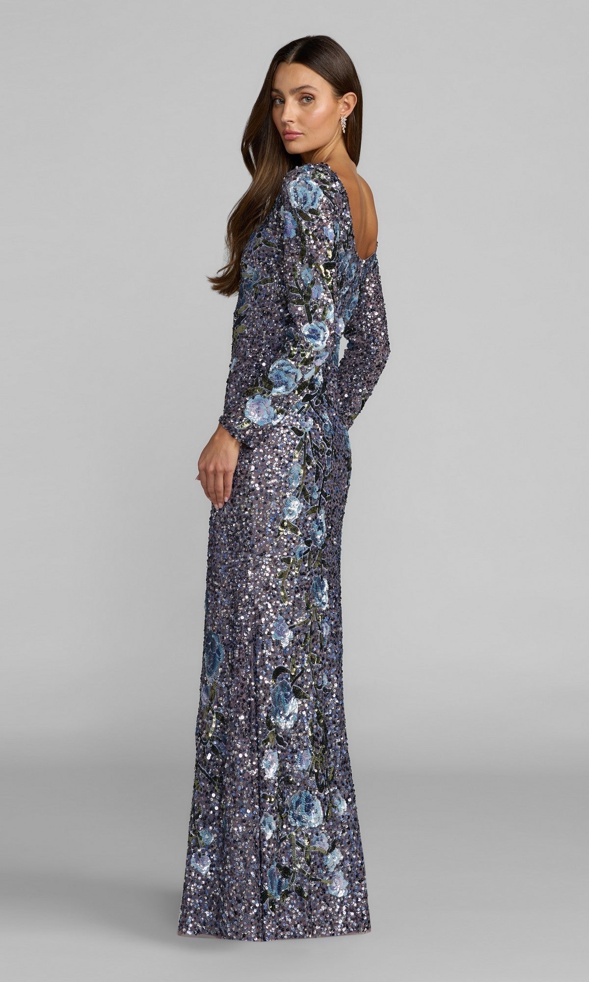 Woman wearing the Mac Duggal 23103 Long Formal Dress, featuring long sleeves, a floor-length silhouette in blue-purple hues with floral patterns and shimmering sequins, standing sideways and looking over her shoulder against a plain light background.