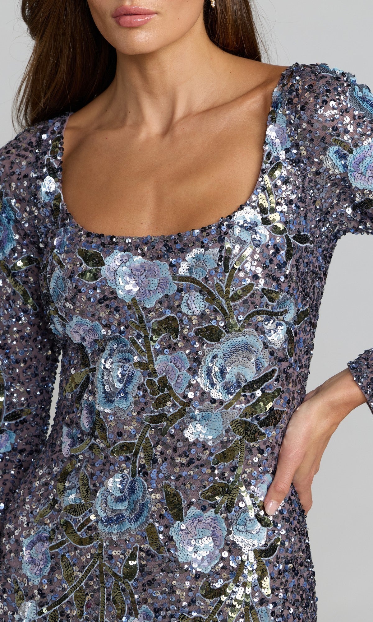 A woman models the Mac Duggal 23103 long formal dress, featuring blue and silver sequin floral embroidery and long sleeves, with one hand resting on her hip.