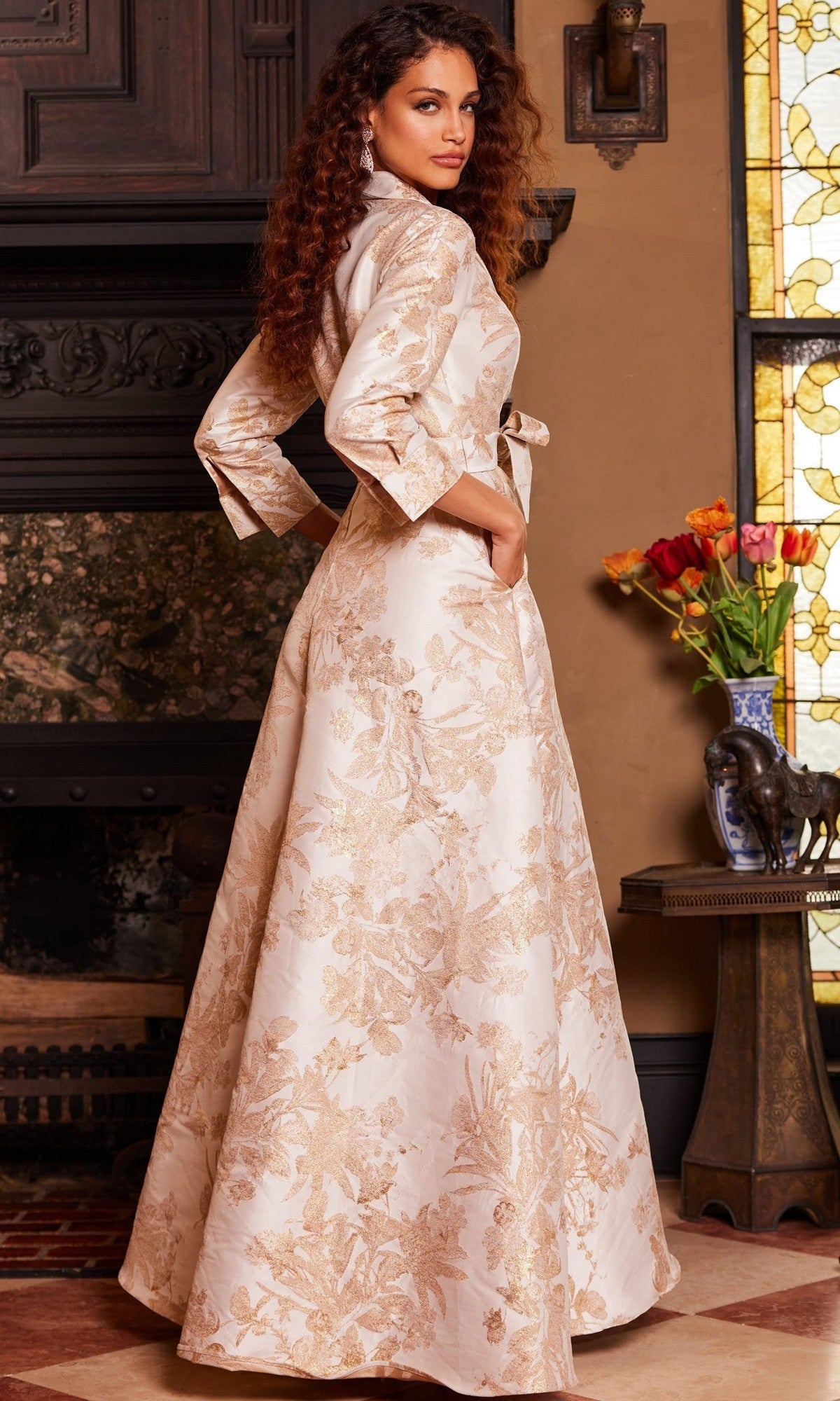 Wearing the Jovani 23178 Long Formal Dress in cream and gold brocade, a woman with long curly hair stands indoors beside a table adorned with flowers, with a stained glass window in the background.