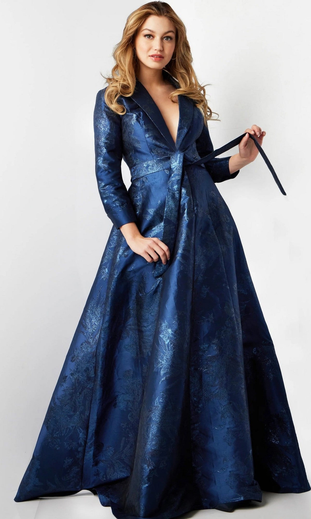 A woman wearing the Long Formal Dress: Jovani 23178, a designer gown featuring a long silhouette, dark blue color, floral pattern, and belted waist, stands against a plain white background.