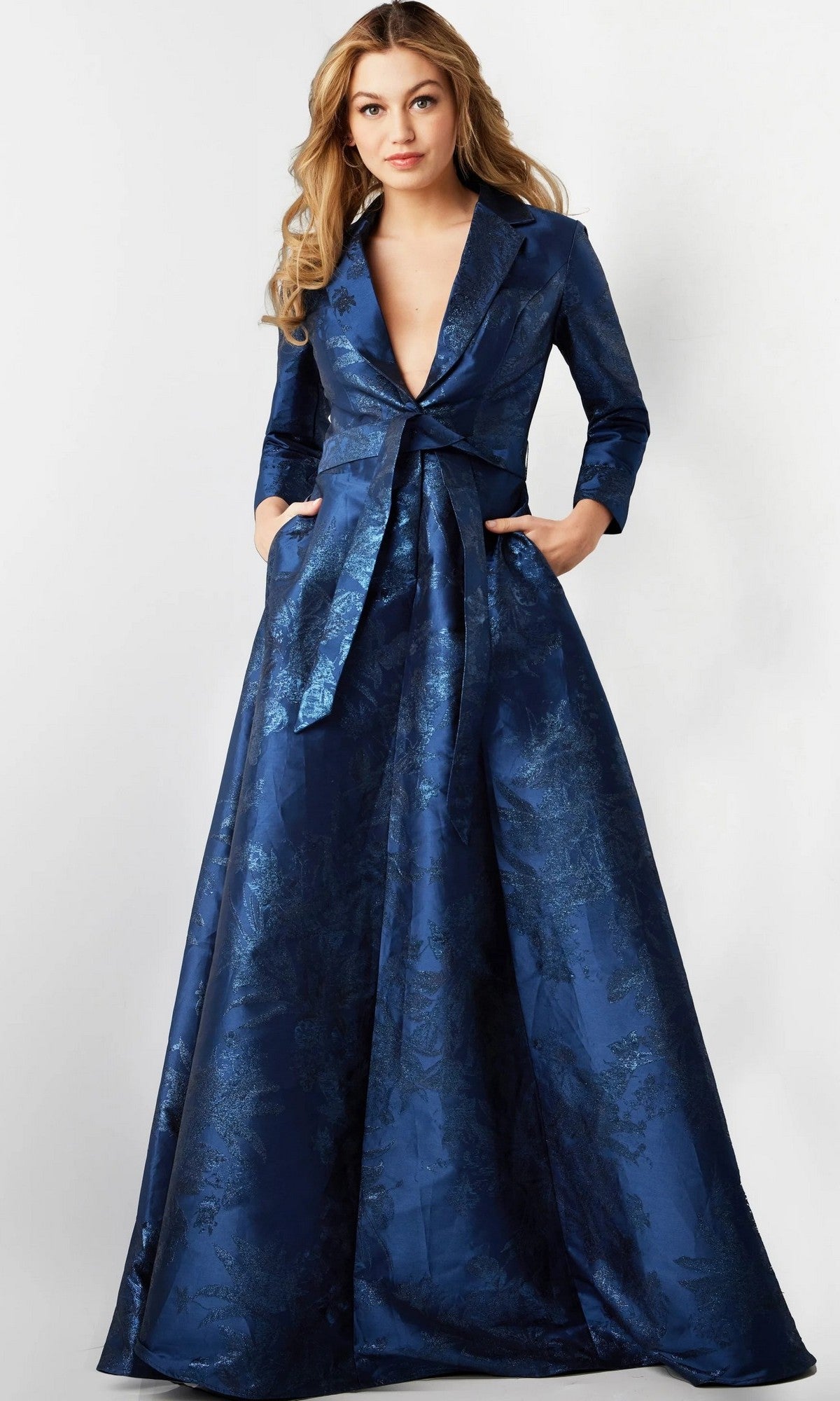 A woman stands against a plain light background, hands in the pockets of the Jovani 23178 Long Formal Dress in navy—a sophisticated designer gown featuring a jacquard fabric, blazer-style top with long sleeves, deep V neckline, and front tie belt—an elegant choice for formal occasions or as a mother-of-the-bride dress.