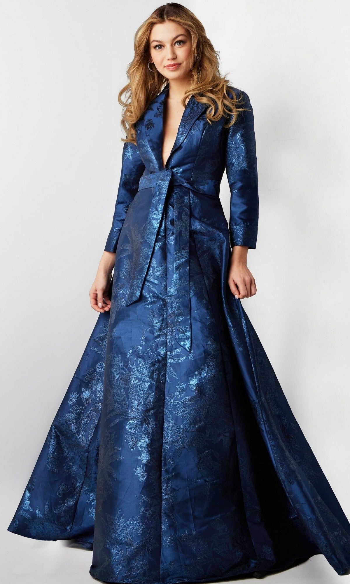 A woman wearing the Long Formal Dress: Jovani 23178—a blue brocade designer gown featuring a belted waist, long sleeves, and a deep V-neckline—stands against a plain background.