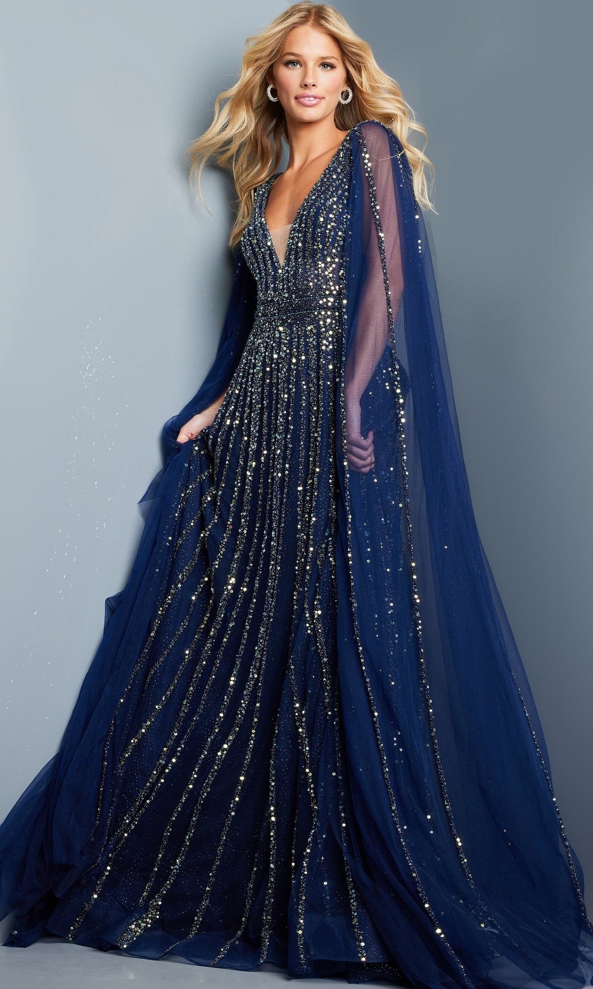 A woman stands against a plain gray background wearing the Jovani 23362 long formal dress in navy, featuring floor-length sheer sleeves and metallic sequin embellishments.