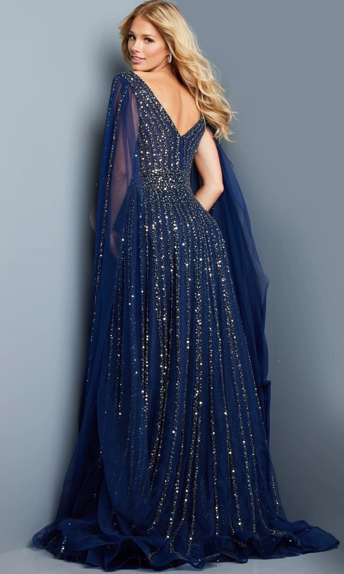 A woman poses with her back and side visible against a plain gray background, wearing the floor-length Jovani 23362 long formal dress—an embellished sequin gown featuring a sheer cape.