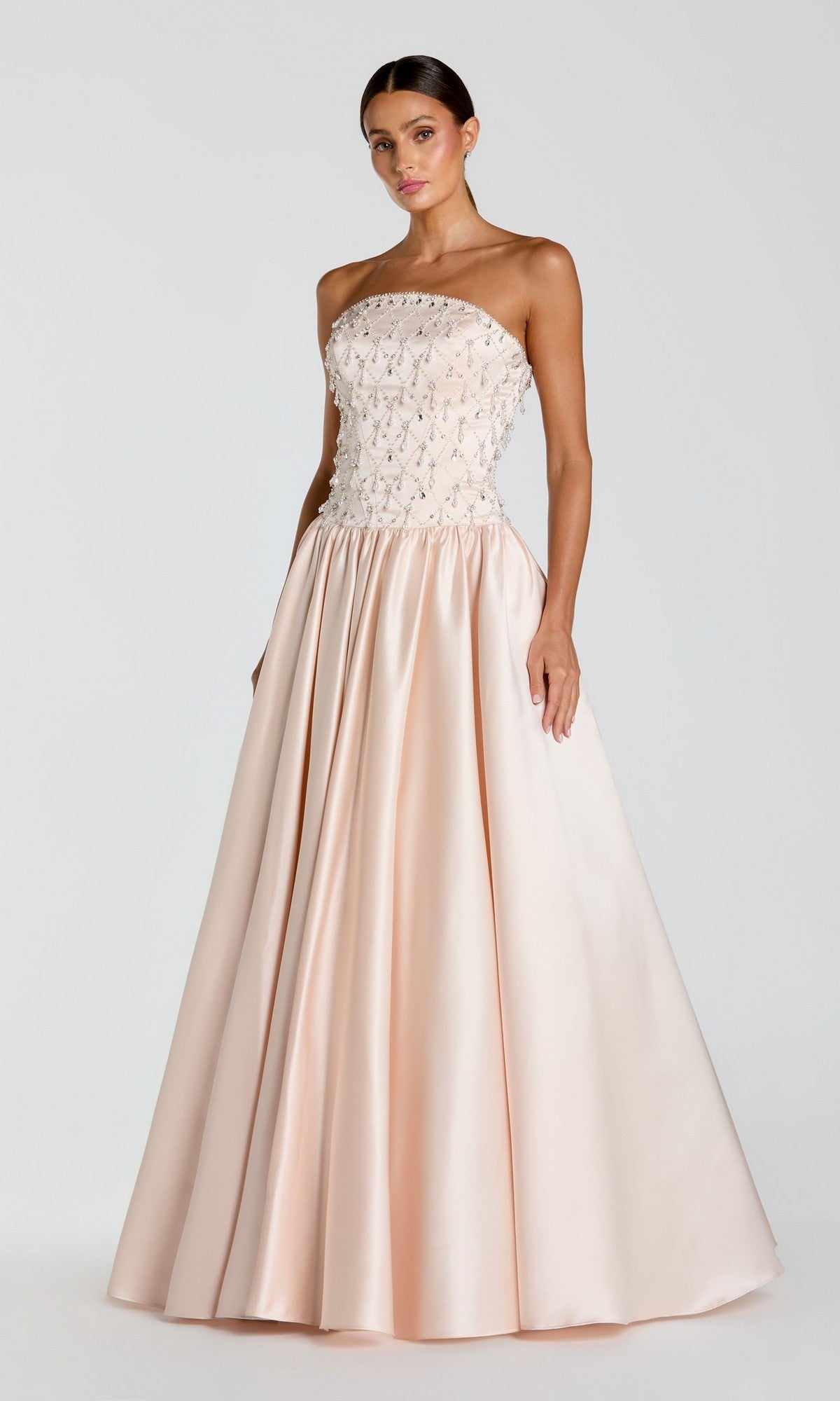 A woman stands wearing the Mac Duggal 2354 Long Formal Dress—a strapless porcelain satin gown with a beaded bodice and a full, floor-length skirt—set against a plain light background.