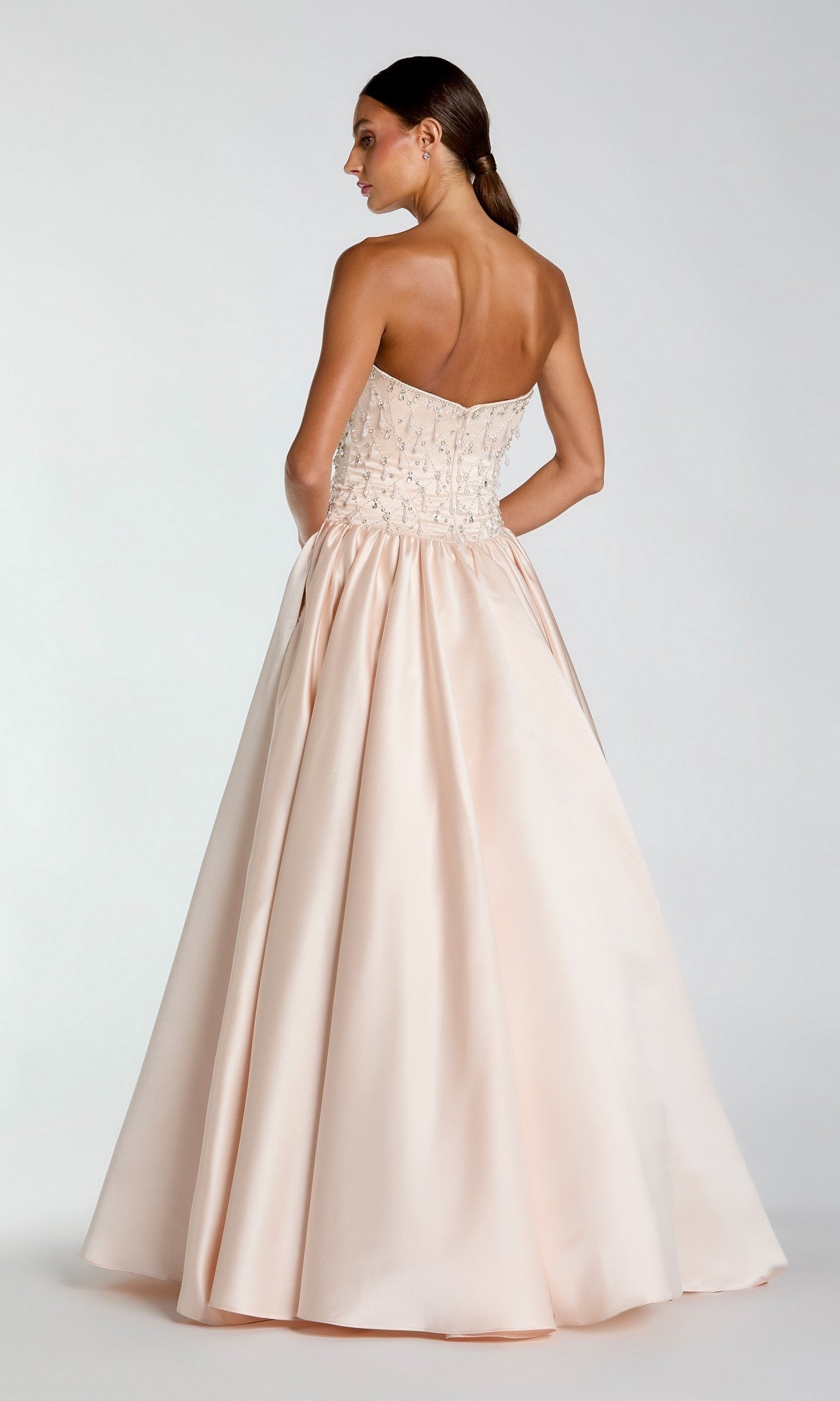 A woman faces away from the camera wearing the Mac Duggal 2354 long formal dress, which features a strapless satin design with a beaded bodice and a full light pink skirt.