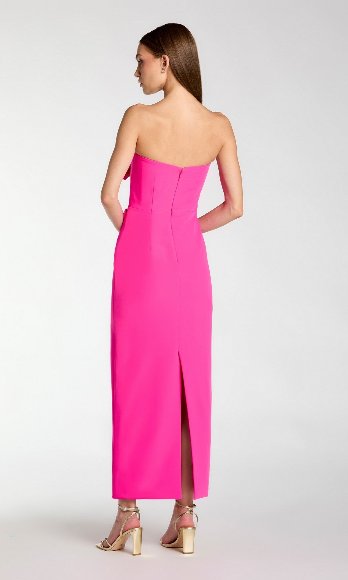 A woman with straight brown hair stands facing away, wearing the Long Formal Dress: Mac Duggal 2361—a bright pink, strapless crepe midi dress featuring a back slit. She completes the look with metallic high-heeled sandals.