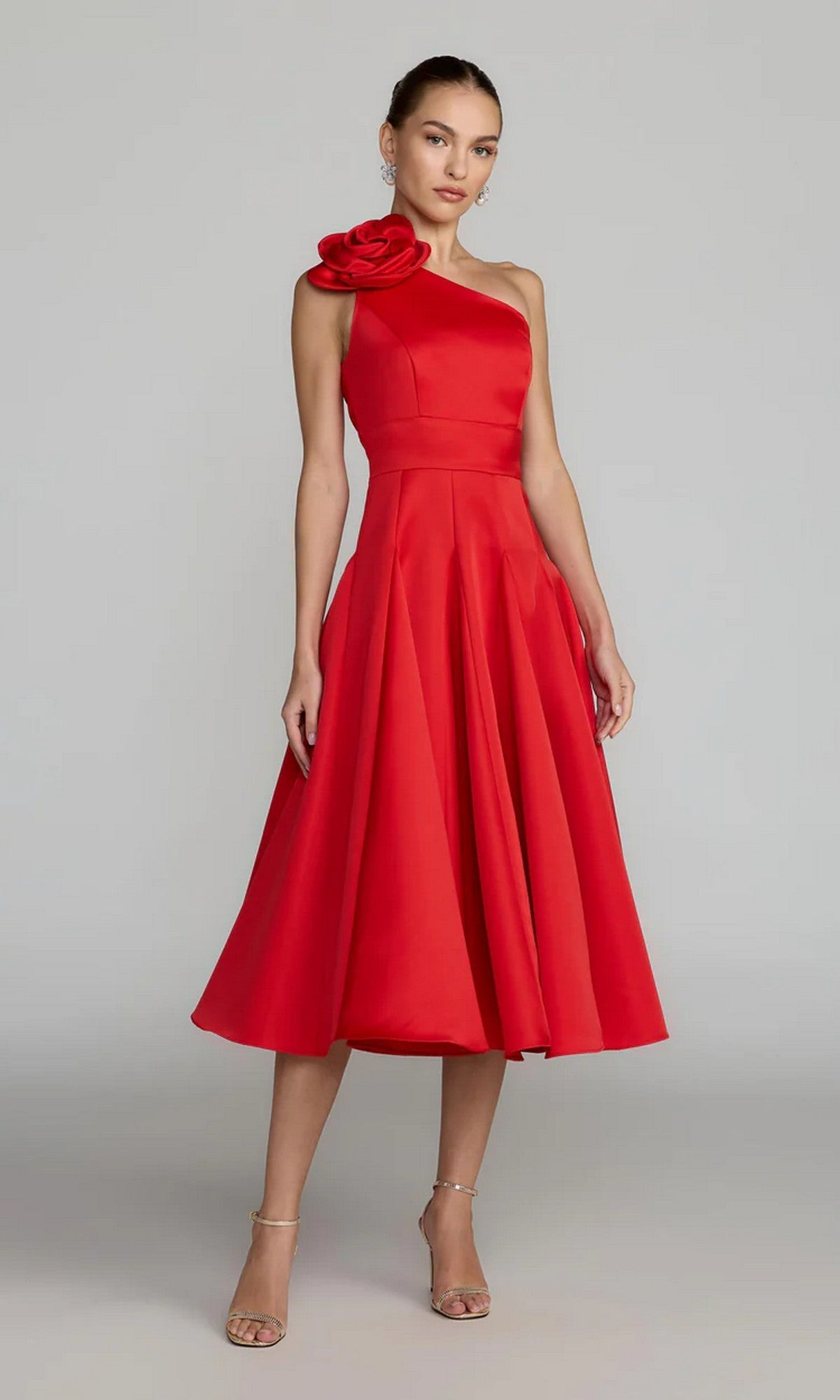 A woman models the Mac Duggal 2364 Midi Party Dress, featuring a bold red one-shoulder design with a statement fabric flower on the shoulder. Crafted from chic satin and falling to the knee, this elegant dress creates a modern silhouette against a plain background.