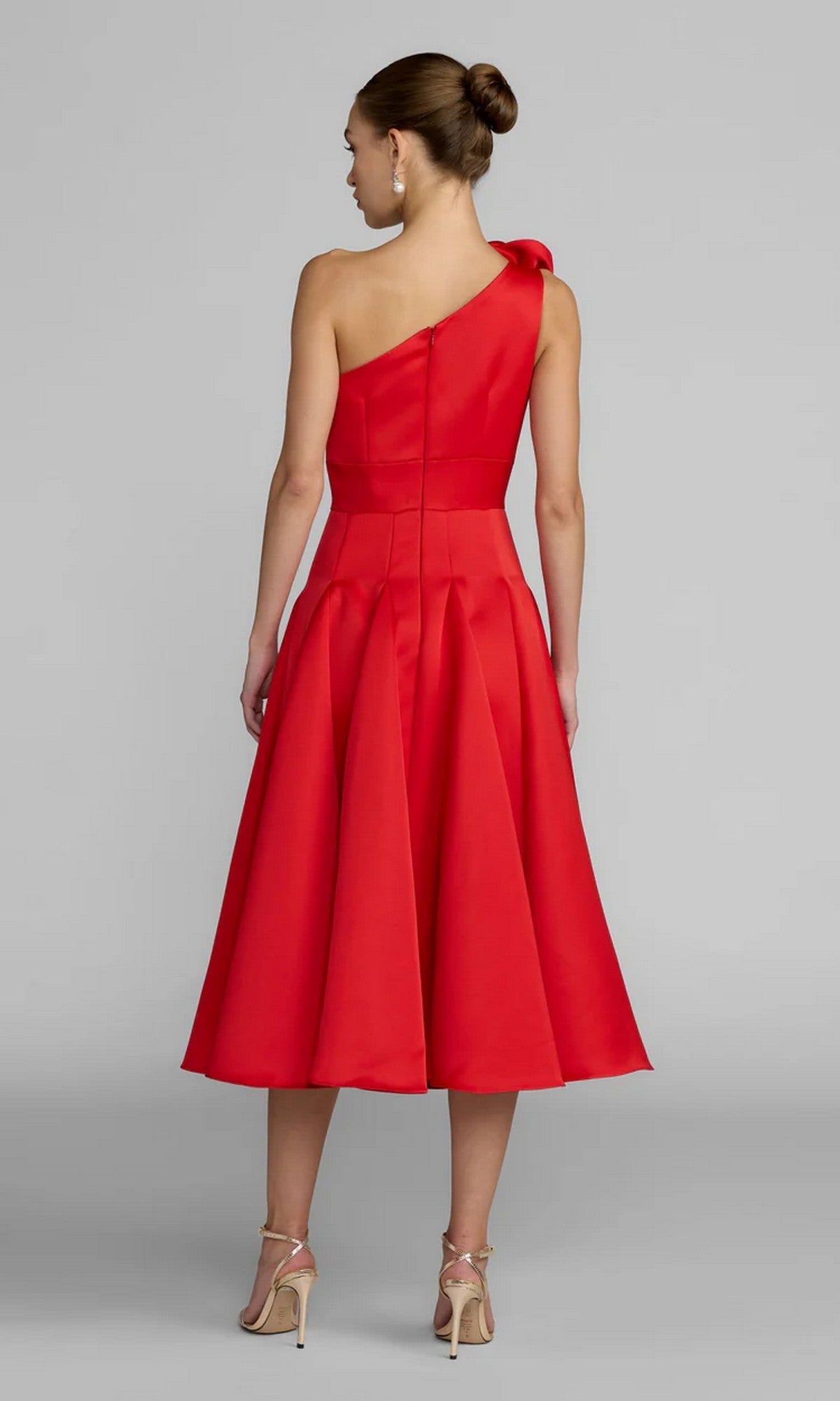 A woman, her hair styled in a bun and beige high-heeled sandals on her feet, stands with her back to the camera wearing the Mac Duggal 2364 Midi Party Dress: a striking red satin, one-shoulder design with elegant pleats and knee-length hem.