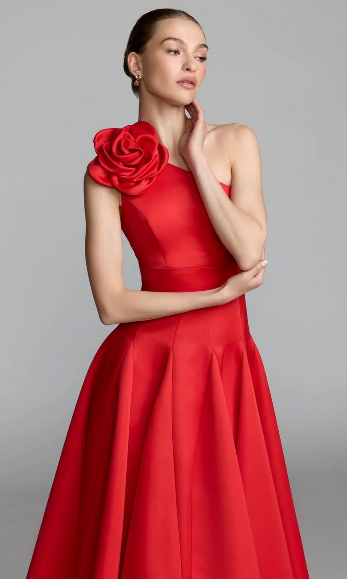 A woman poses against a plain background with one hand near her face, wearing the Mac Duggal 2364 Midi Party Dress—a sleeveless red dress featuring an asymmetrical neckline and a large rose detail on one shoulder.