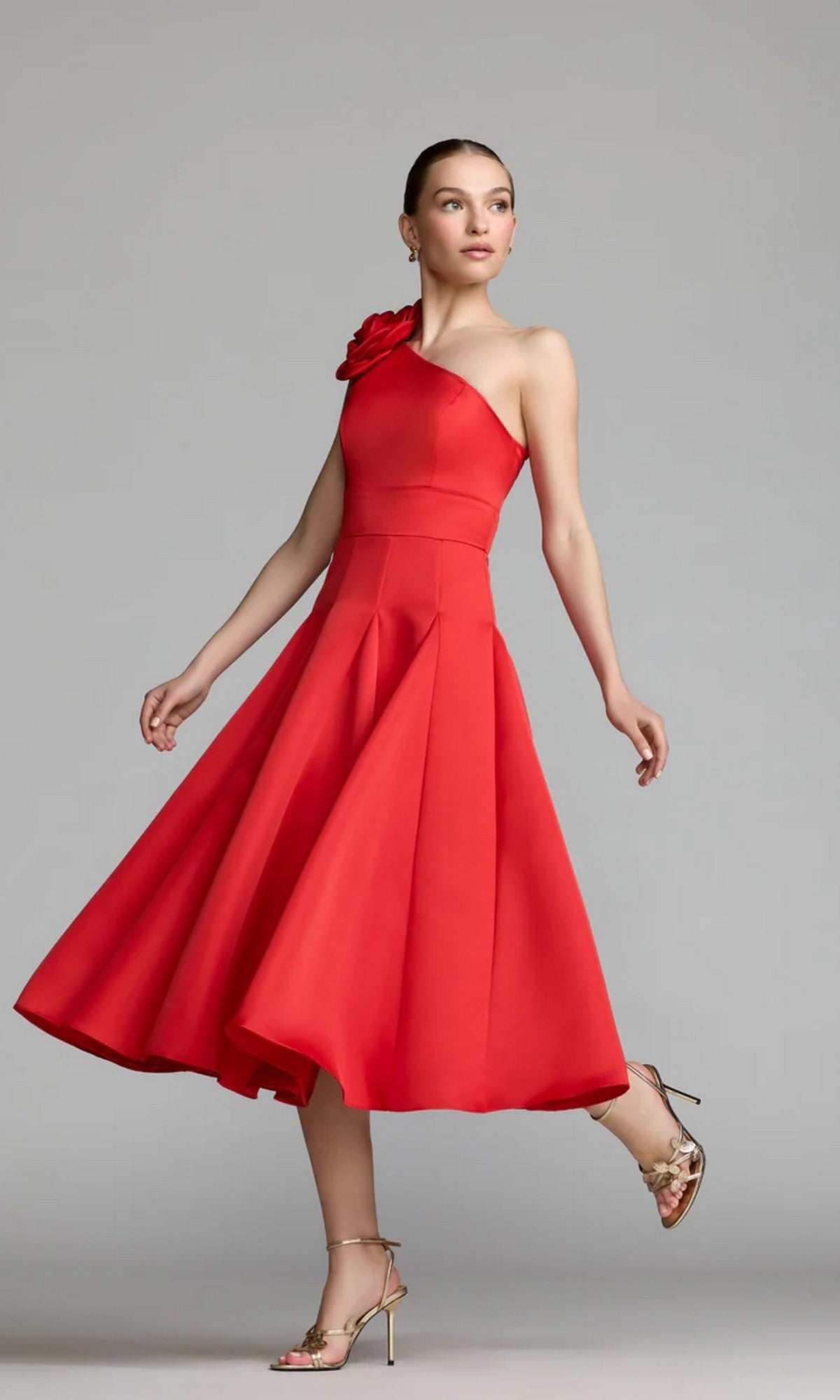 A woman models the Mac Duggal 2364 midi party dress in bold red—a striking one-shoulder satin dress with a floral accent at the strap—paired with gold high-heeled sandals, standing against a plain gray background.