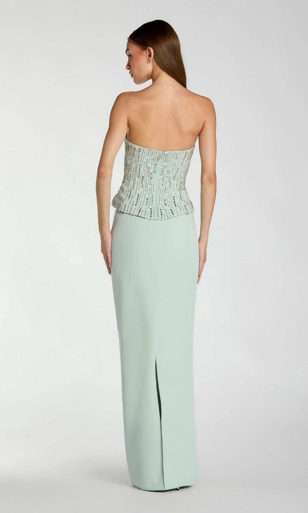 A woman stands with her back to the camera against a plain background, wearing the Long Formal Dress: Mac Duggal 2366—an elegant mint strapless gown featuring a beaded bodice and a long, light green crepe skirt with a back slit.