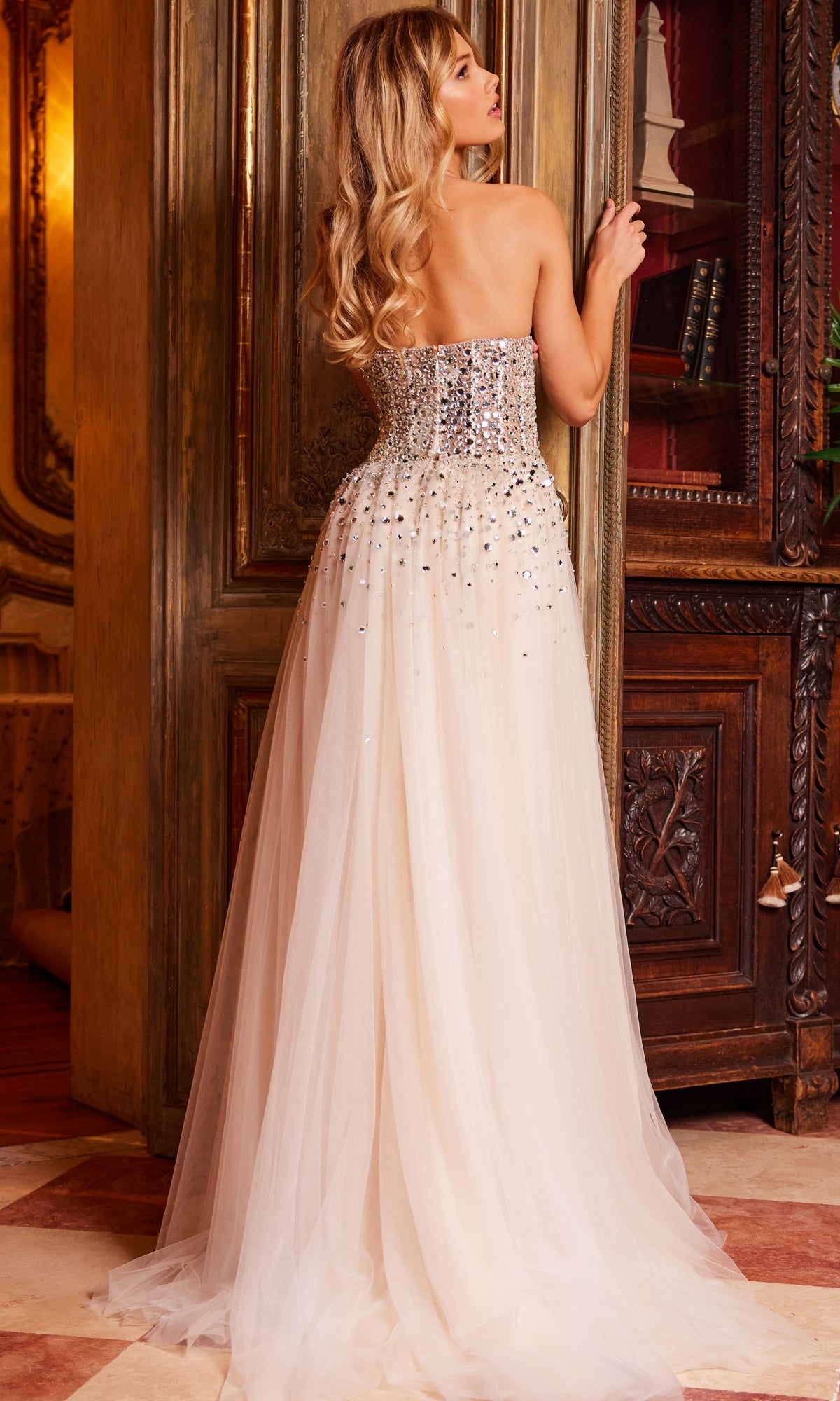 A woman with long blonde hair, seen from behind in an ornate room, wears the Jovani Strapless Beaded Nude Prom Ball Gown 23712 featuring a beaded bodice and floor-length tulle skirt.