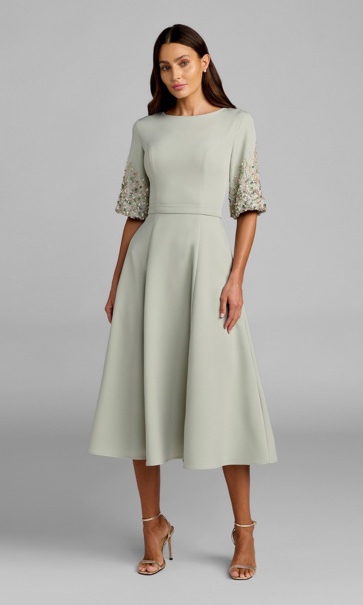 A woman wearing a sage Tea-Length Party Dress (Mac Duggal 2372) with floral-embellished half sleeves and beige high-heeled sandals, standing against a plain gray background.