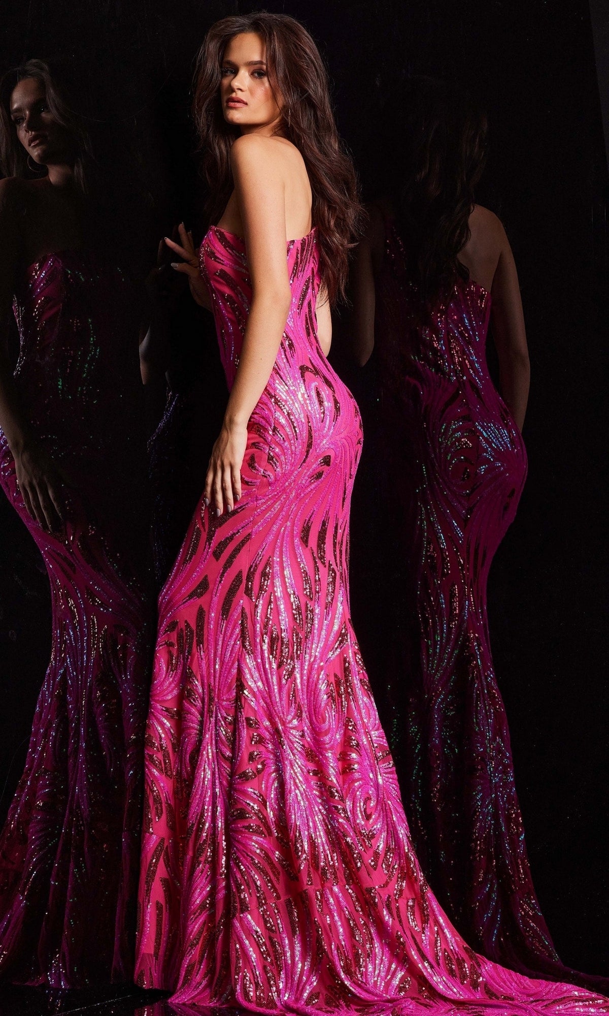 A woman models the Jovani One-Shoulder Sequin Long Prom Dress 23876 in bright pink, posing by a mirror to display its sparkling details and elegant floor-length silhouette against a dark background.