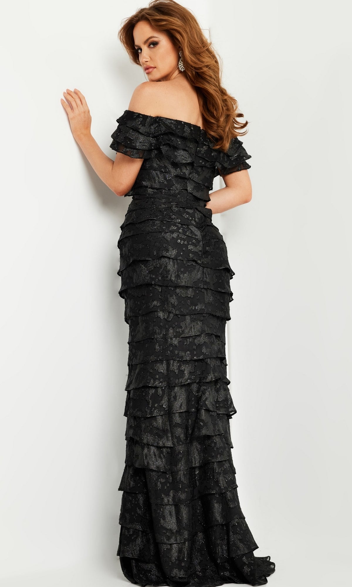 A woman stands with her back to the camera, looking over her shoulder in the Jovani 23890 Long Black Formal Dress, featuring off-the-shoulder tiered ruffles, set against a plain white background.