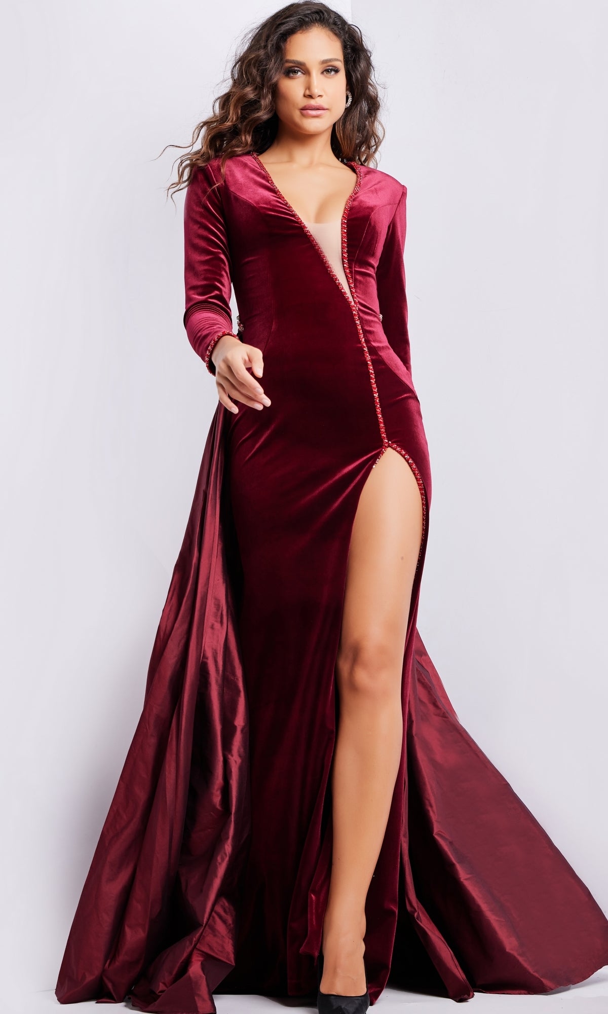 A woman stands against a plain background wearing the Jovani Long Sleeve Long Velvet Formal Dress 23943, featuring a fitted bodice, high slit, and deep red color.