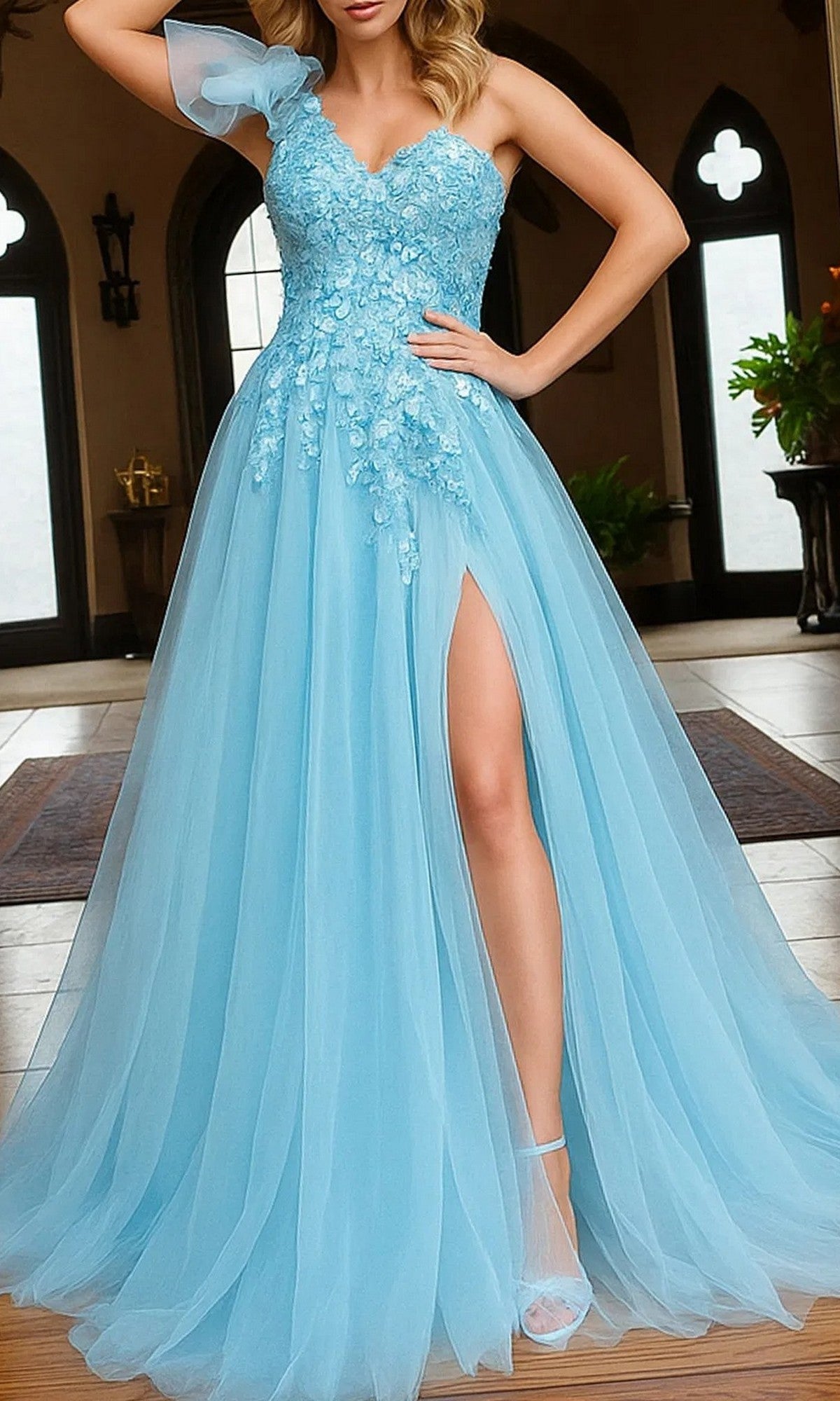 A woman wears the Long Prom Dress: Jovani 23951 in Blue/Multi with floral embroidery, a sheer one-shoulder strap, and a high leg slit, standing indoors before arched windows.