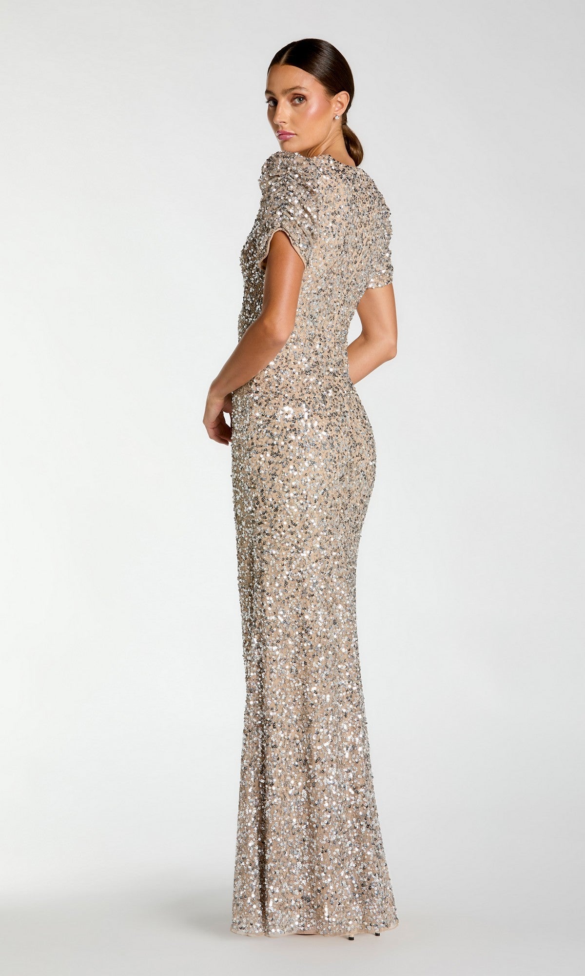 A woman faces away, modeling the Mac Duggal 24003—a shimmering floor-length silver short sleeve sequin formal dress—set against a light plain background.