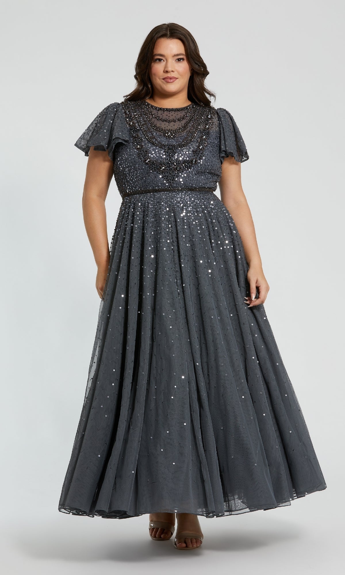 A woman wears the Plus-Size Charcoal Formal Dress: Mac Duggal 24025, a floor-length, short-sleeve evening gown embellished with sequins and beading, standing against a plain light background.