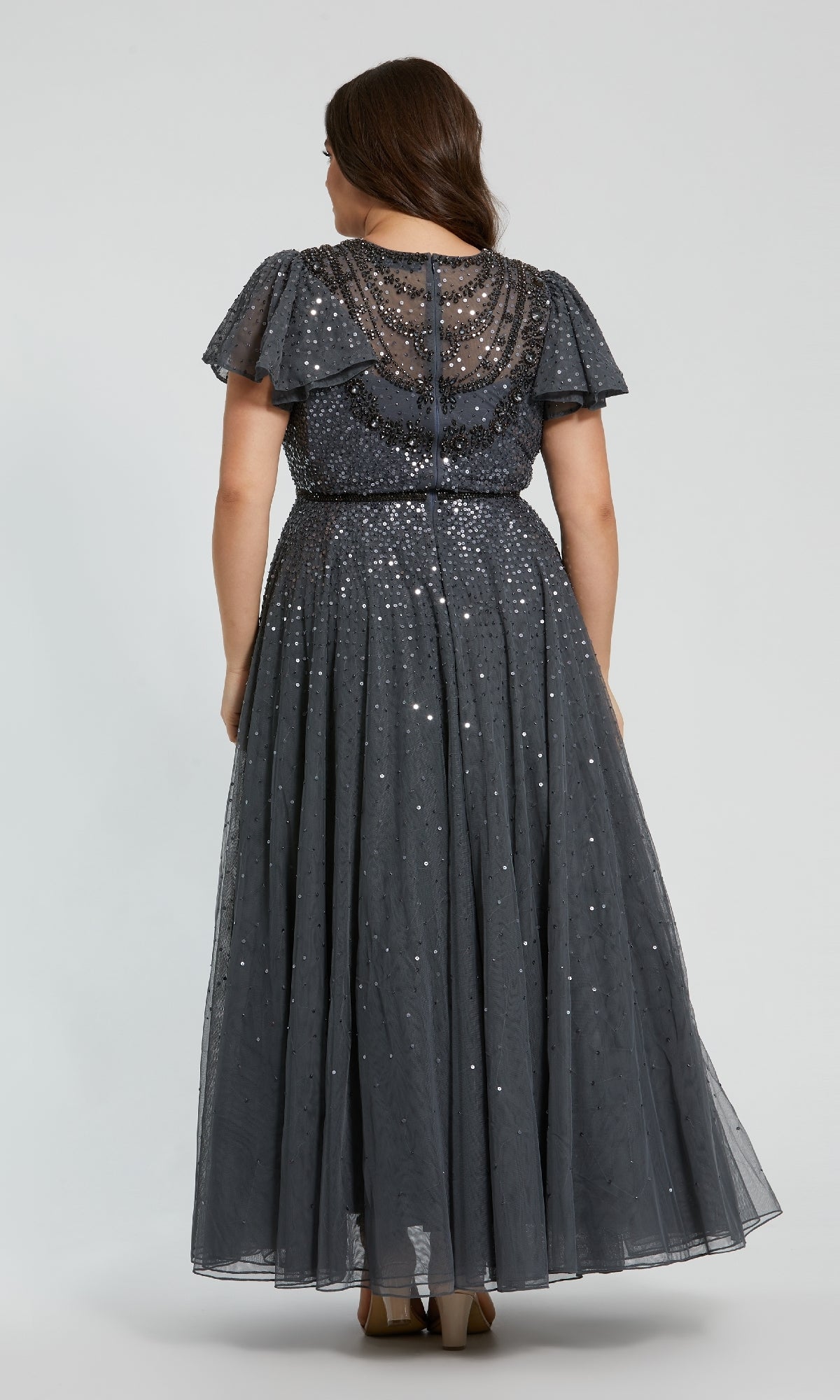 A woman is photographed from behind in a Mac Duggal 24025 Plus-Size Charcoal Formal Dress, an ankle-length, short-sleeved sequin evening gown, standing against a plain light background.