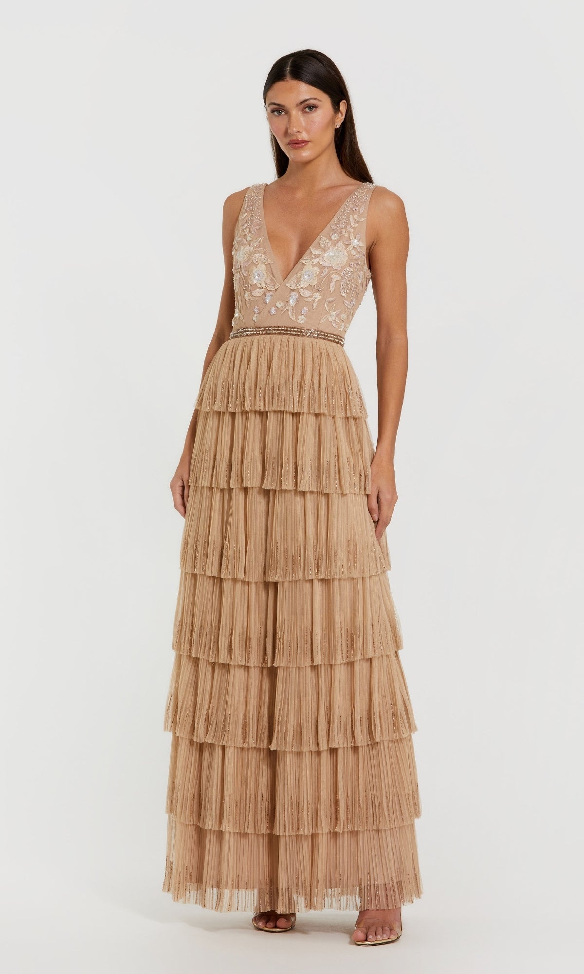 A woman stands against a white background wearing the Mac Duggal 24061: a sleeveless, floor-length nude formal dress with a sheer bead-embellished bodice and tiered ruffled skirt.
