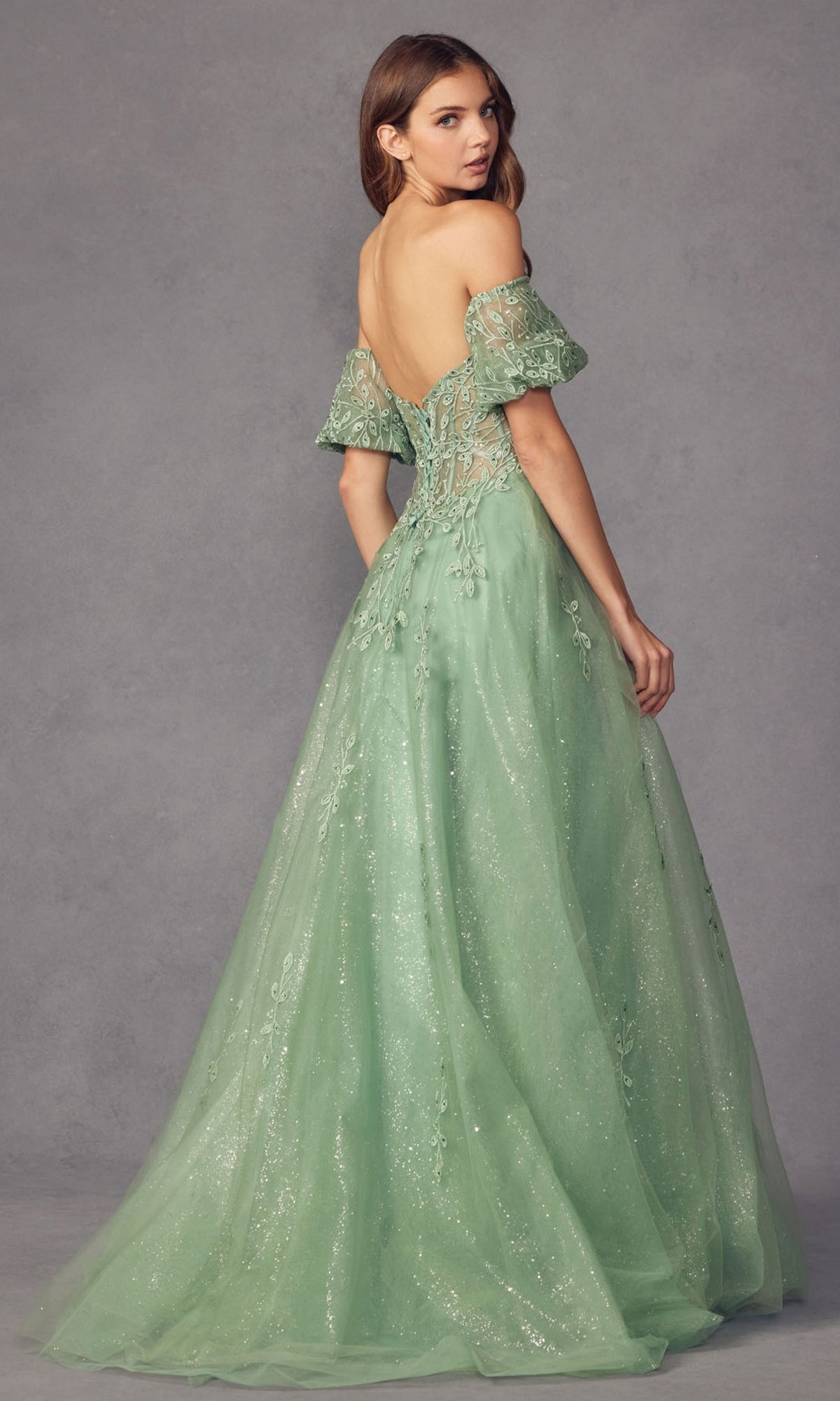 A woman in the Puff-Sleeve Strapless Glitter Prom Ball Gown 2409, a light green, floor-length dress with lace, sequins, and detachable puff sleeves, stands facing backward against a gray backdrop.