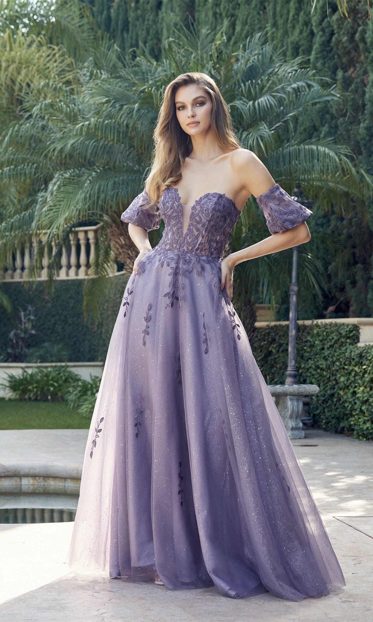 A woman poses outdoors in the Puff-Sleeve Strapless Glitter Prom Ball Gown 2409, featuring floral embroidery and a sheer, flowing skirt. Lush greenery and a pool in the background highlight her elegant style.